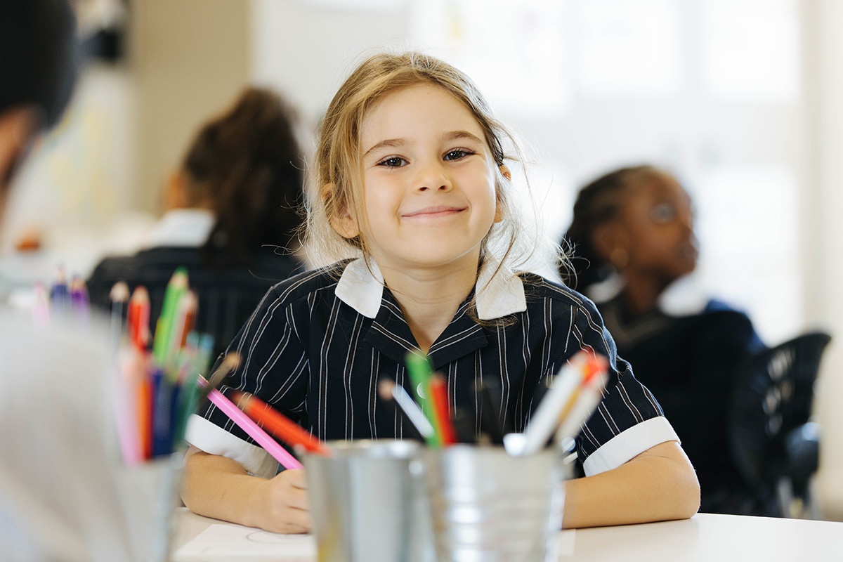 Smiling primary aged girl sitting at desk with small bucket of pencils