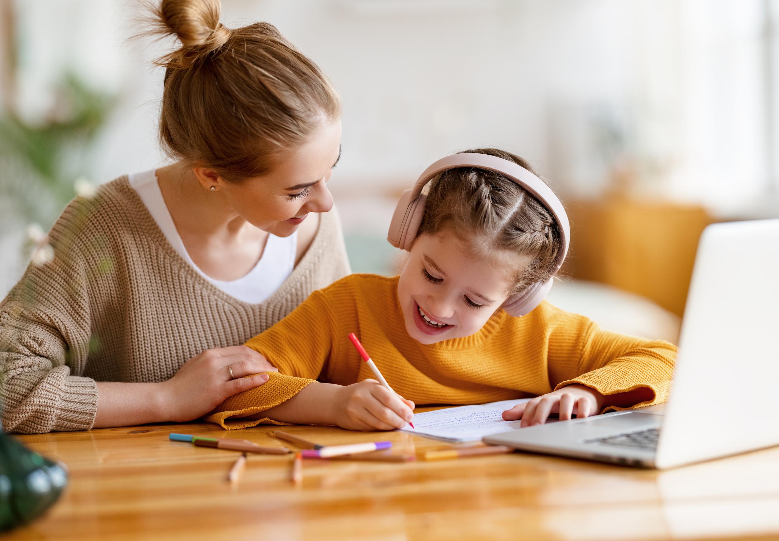 A child wearing headphones smiles while writing with colored pencils, assisted by an adult in a cozy indoor