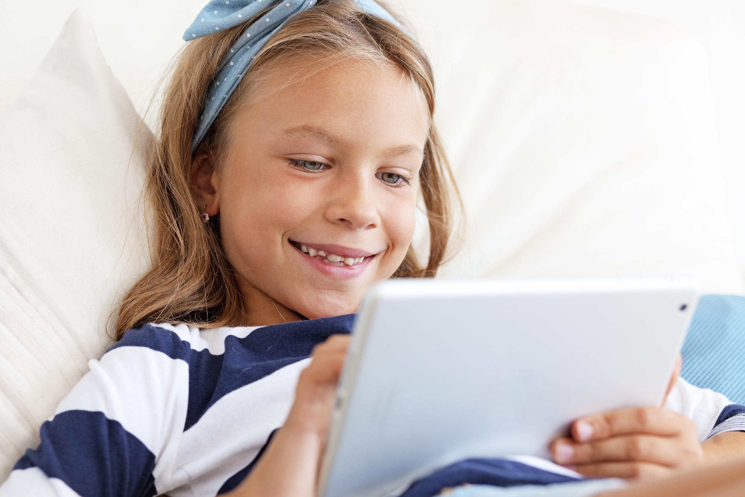 A girl with long hair and a blue headband smiles while using a tablet, lying on a couch.