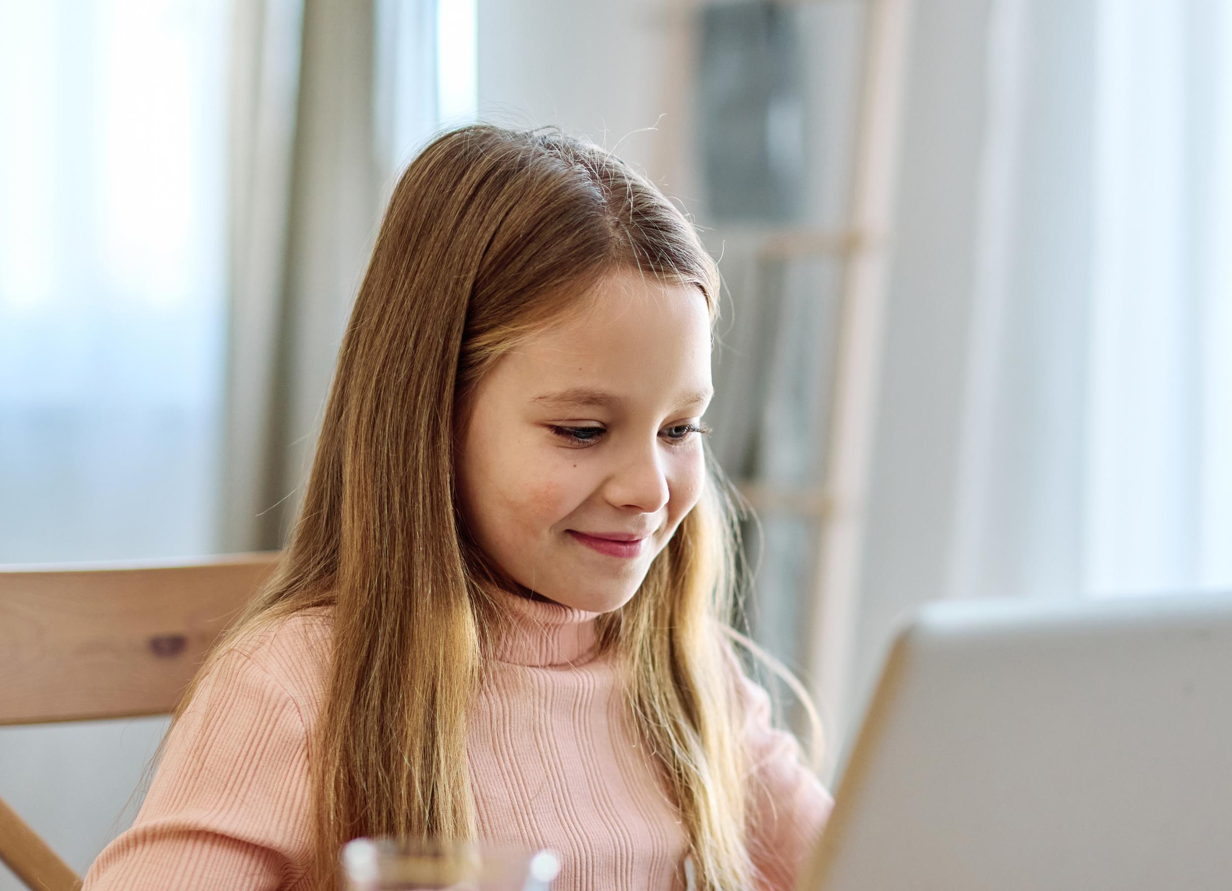 A girl with long hair smiles while looking at a laptop screen, seated at a wooden table
