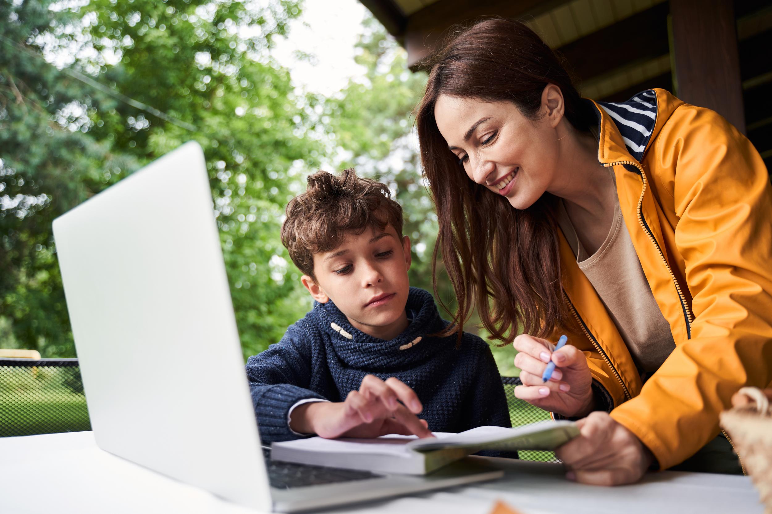 A mother assists her son with schoolwork on a laptop outdoors at ACC Moreton, surrounded by greenery.