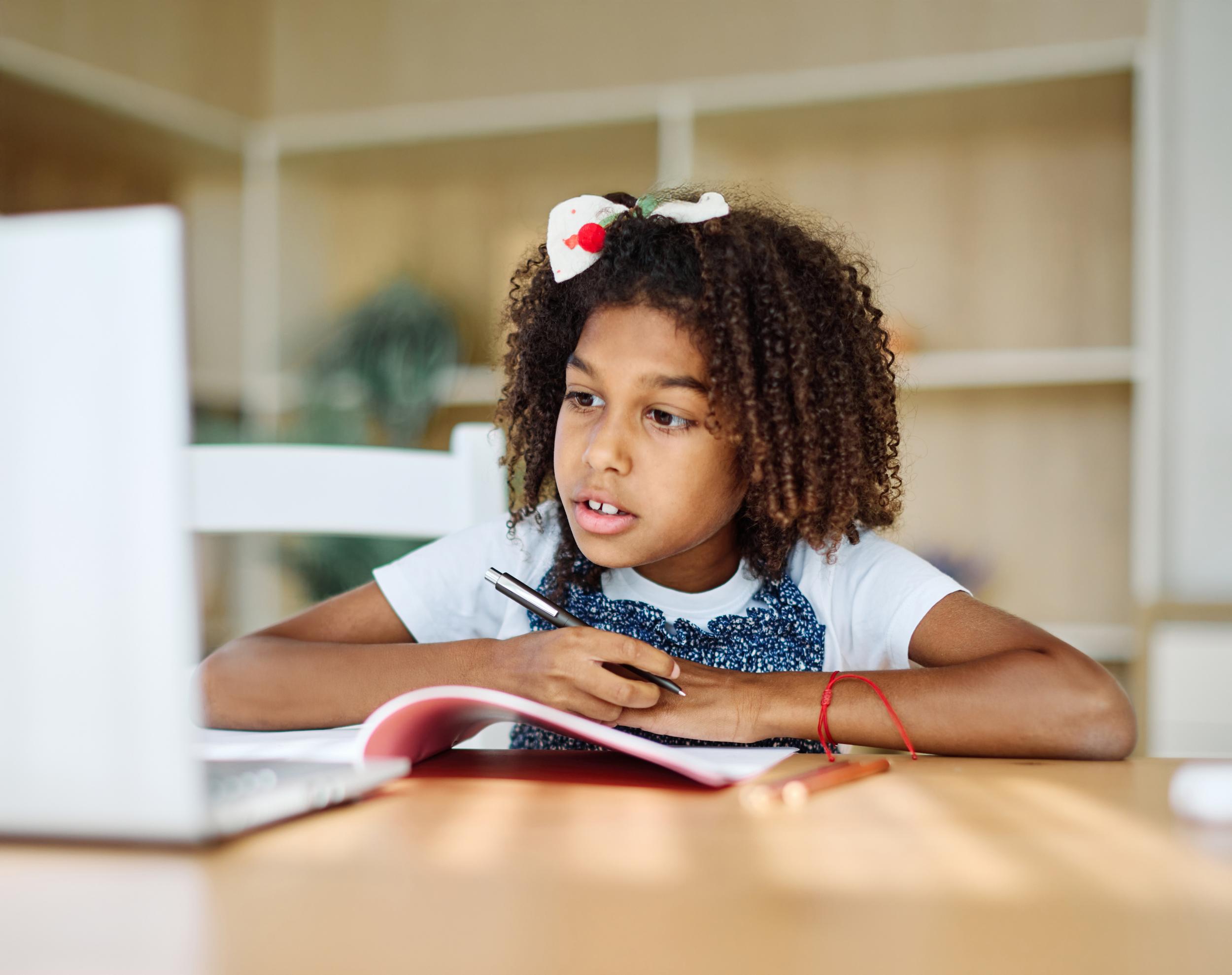 A student with curly hair sits at a desk, focused on a laptop, holding a pen and looking