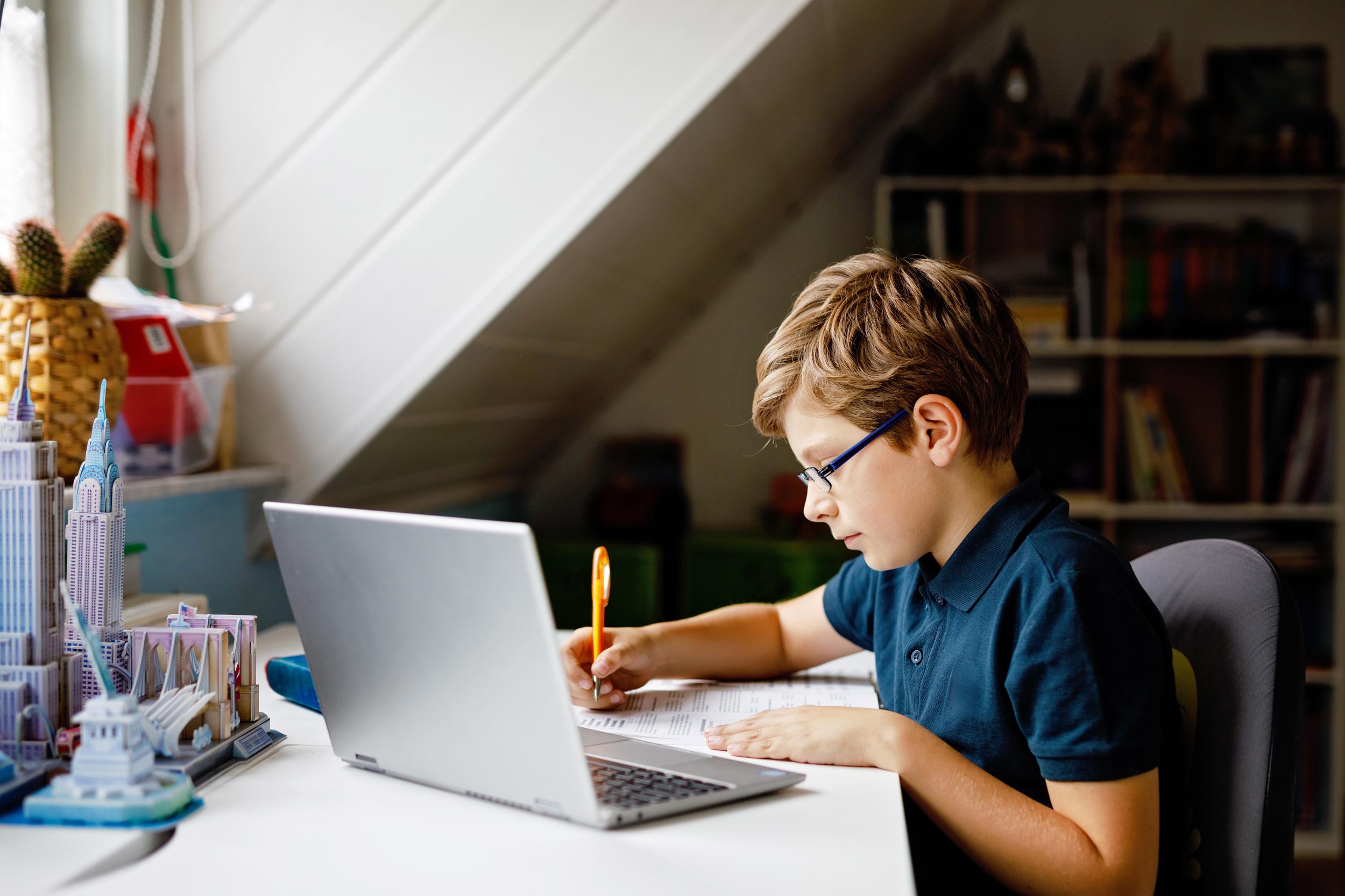 A student with glasses sits at a desk, writing in a notebook while using a laptop. Bookshelves are