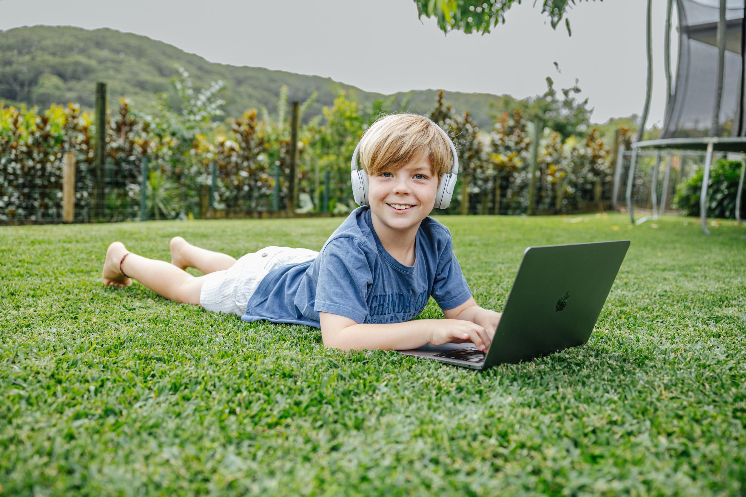 A student at ACC Moreton lies on the grass, wearing headphones and using a laptop, engaged in learning