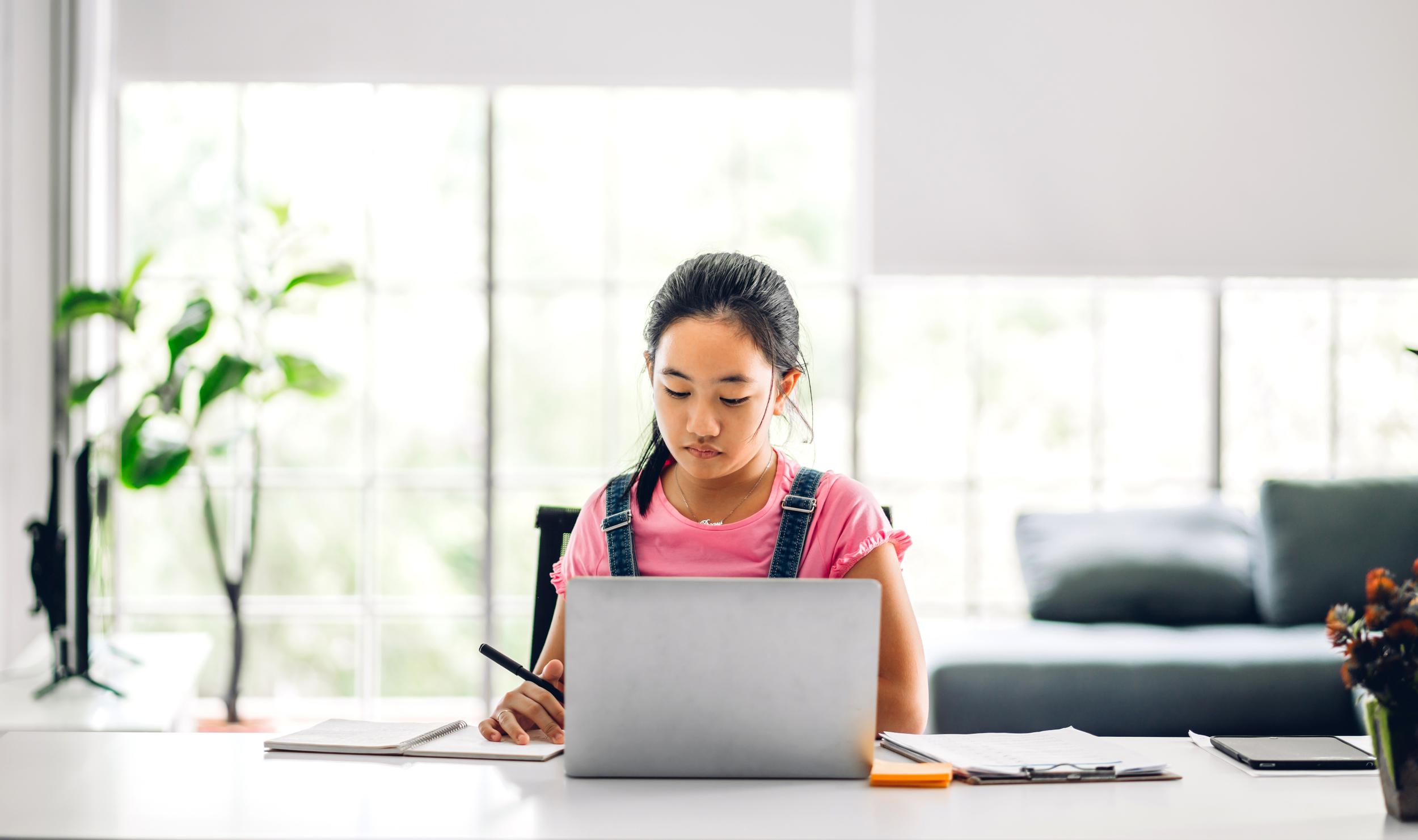 A student sits at a desk, focused on a laptop, writing notes with a pen. Natural light fills
