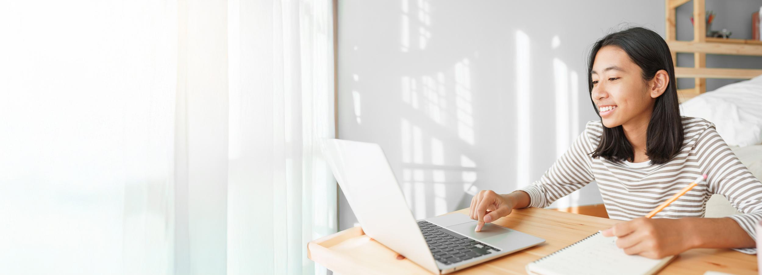 A student at ACC Moreton sits at a desk, smiling while using a laptop, with a notebook