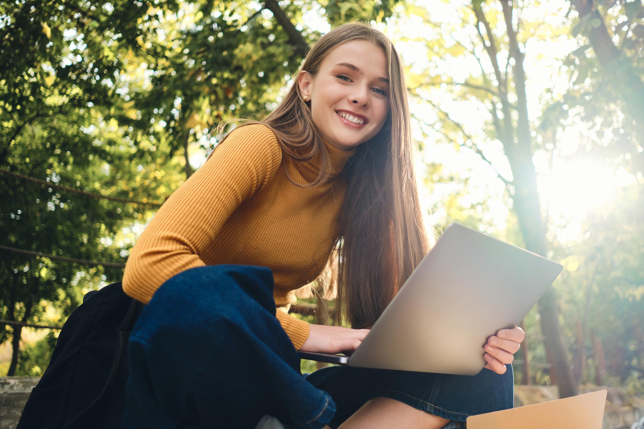 A student sits outdoors on a sunny day, smiling while using a laptop. Trees and sunlight create
