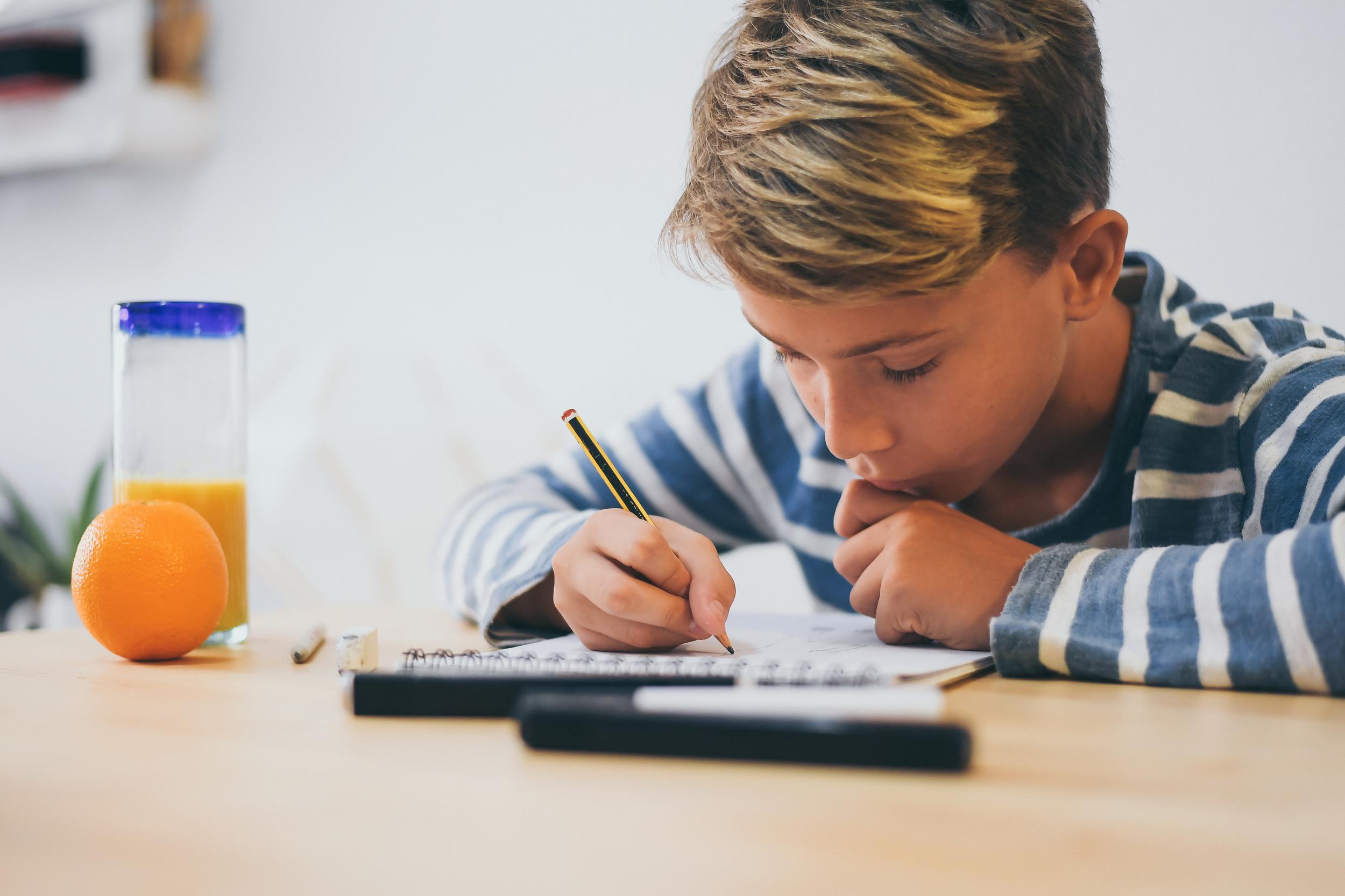 A student sits at a table, focused on writing in a notebook, with a pencil in hand.