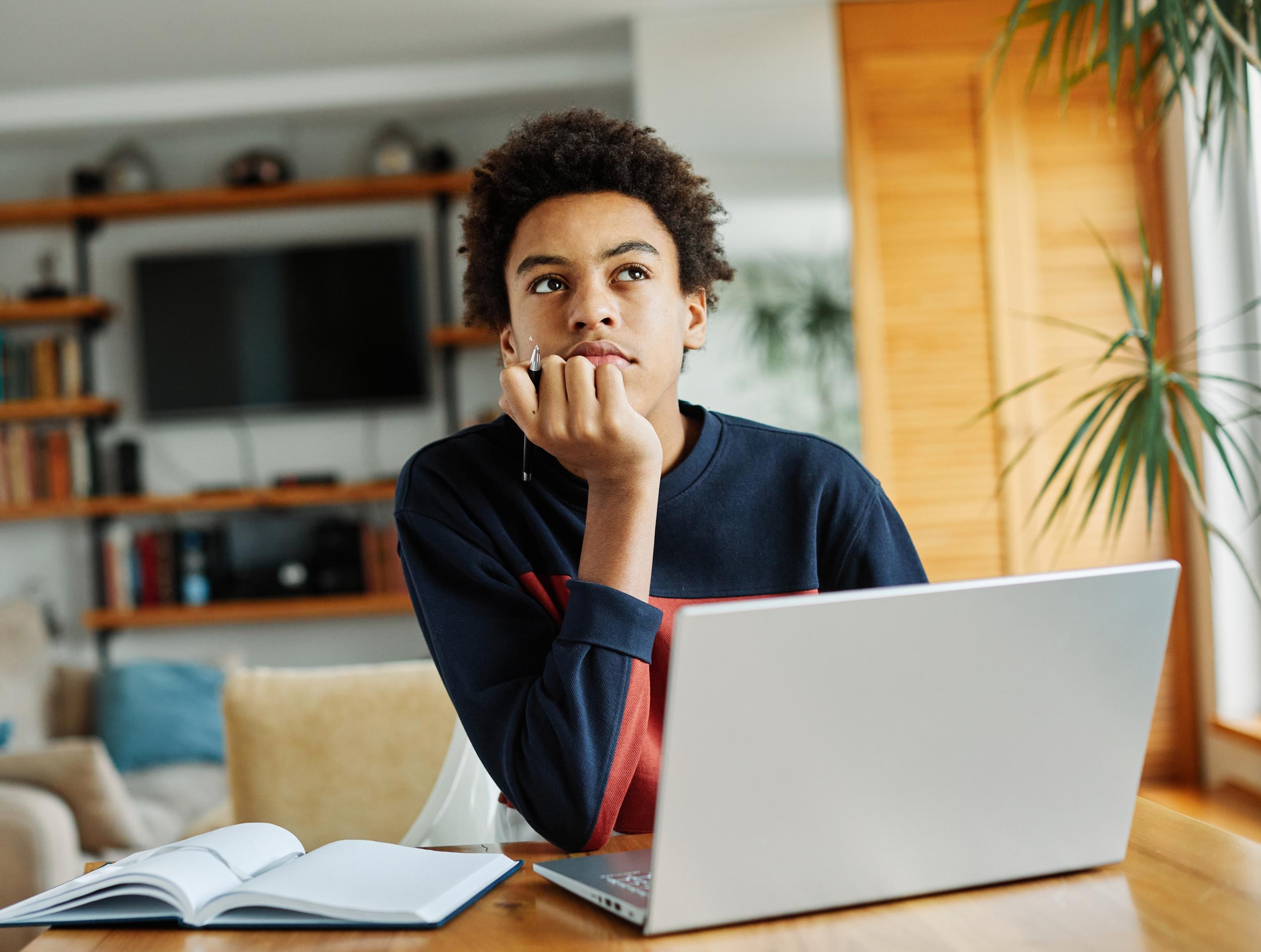 Student sitting at a table, looking thoughtfully at a laptop, with an open notebook beside him