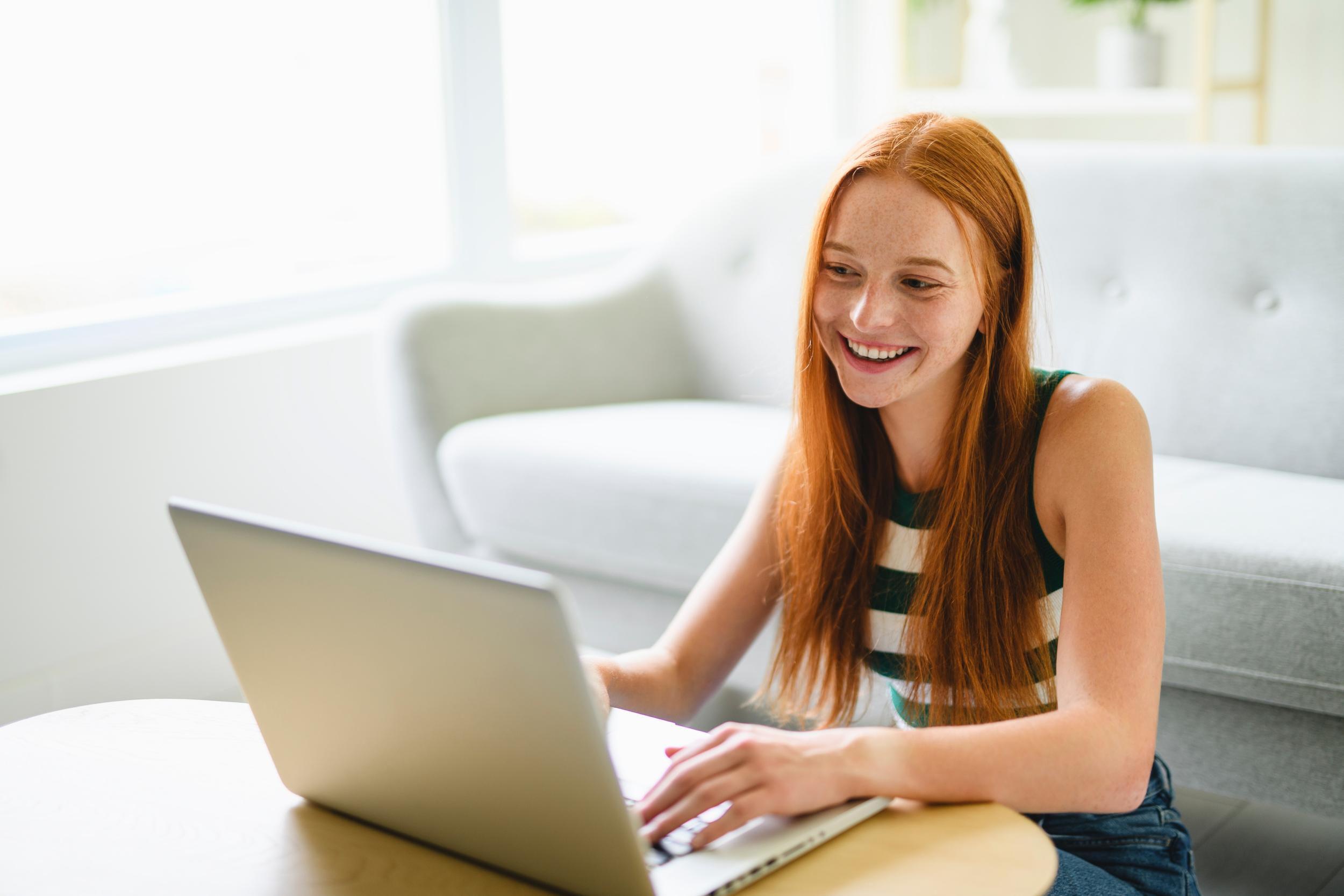 A student at ACC Moreton smiles while working on a laptop at a table in a bright, modern