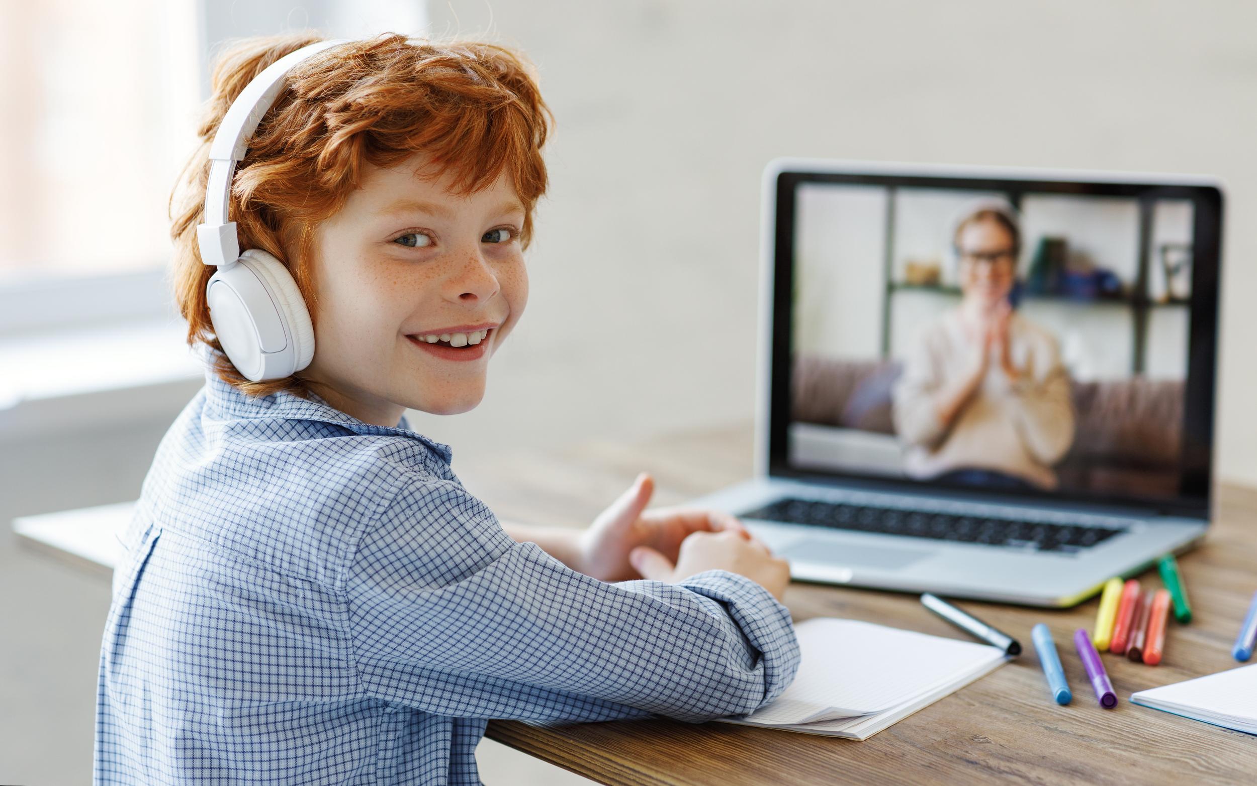 A student wearing headphones smiles while sitting at a desk, engaging in an online lesson on a laptop.