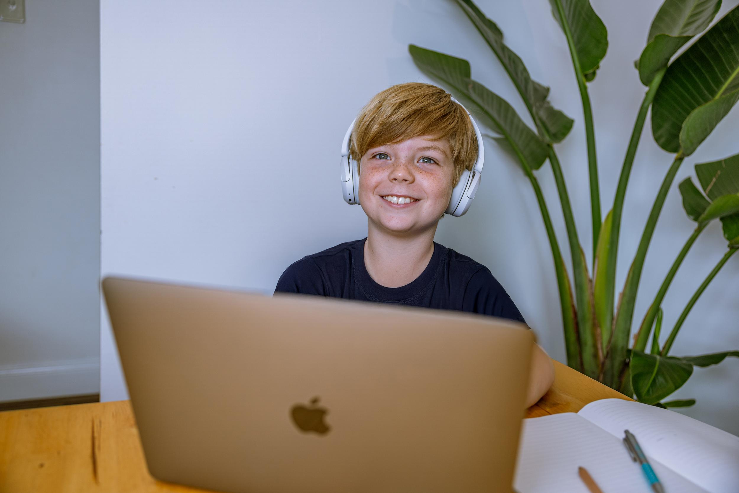 A student wearing headphones smiles while sitting at a table with a laptop and notebook, surrounded by