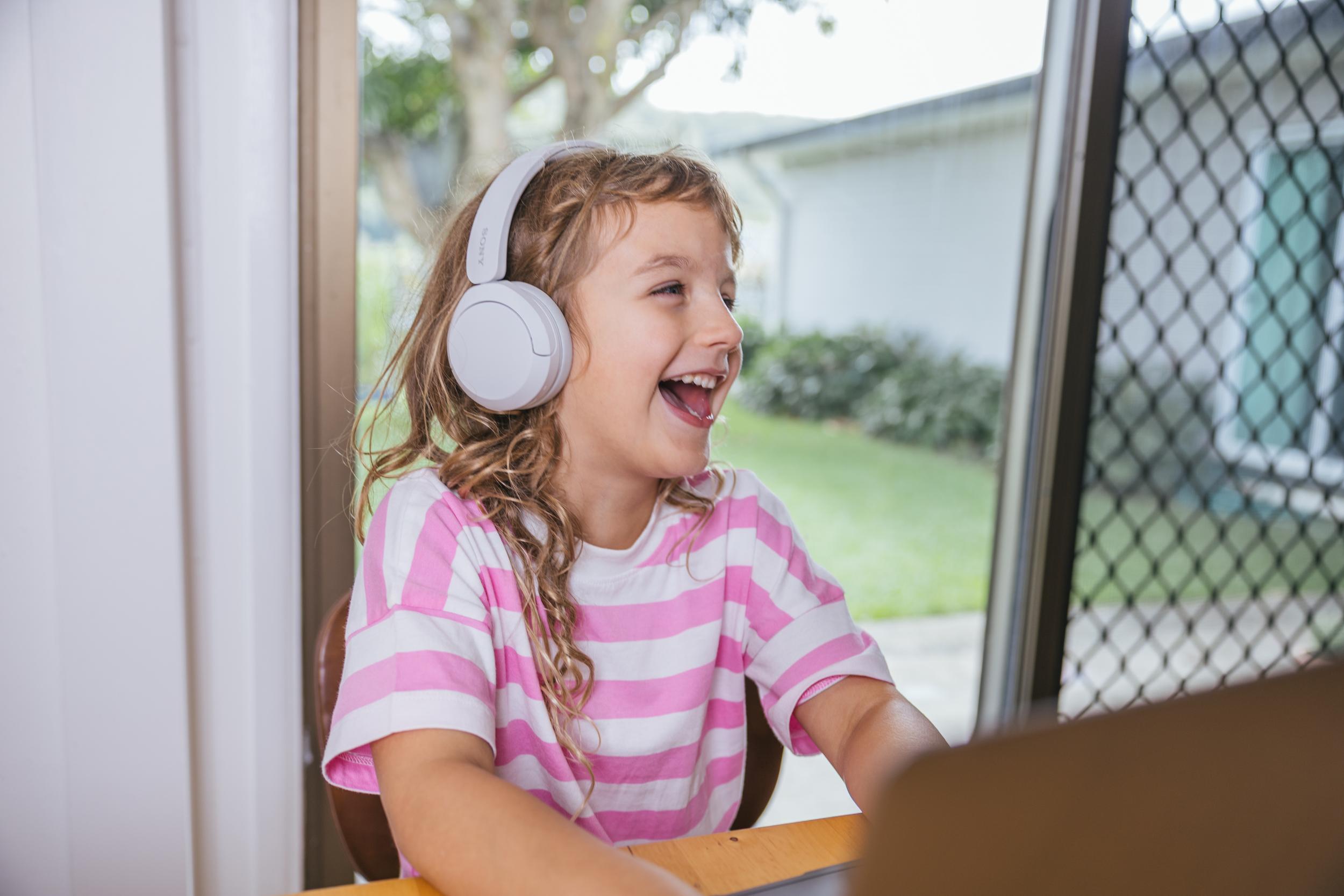 A student wearing headphones smiles while engaging with a laptop at a desk near a window.