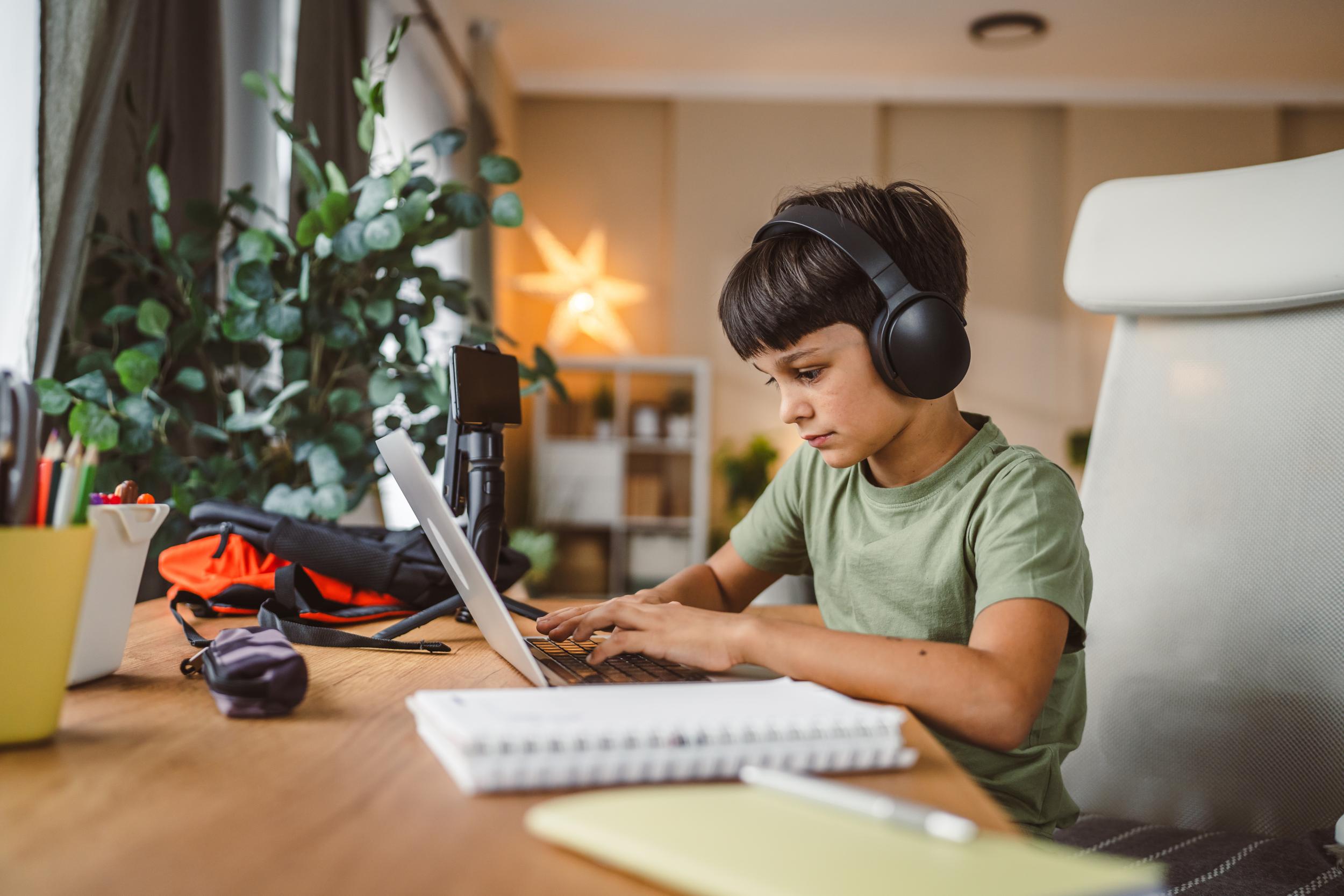 A student wearing headphones sits at a desk, focused on a laptop, with stationery and plants