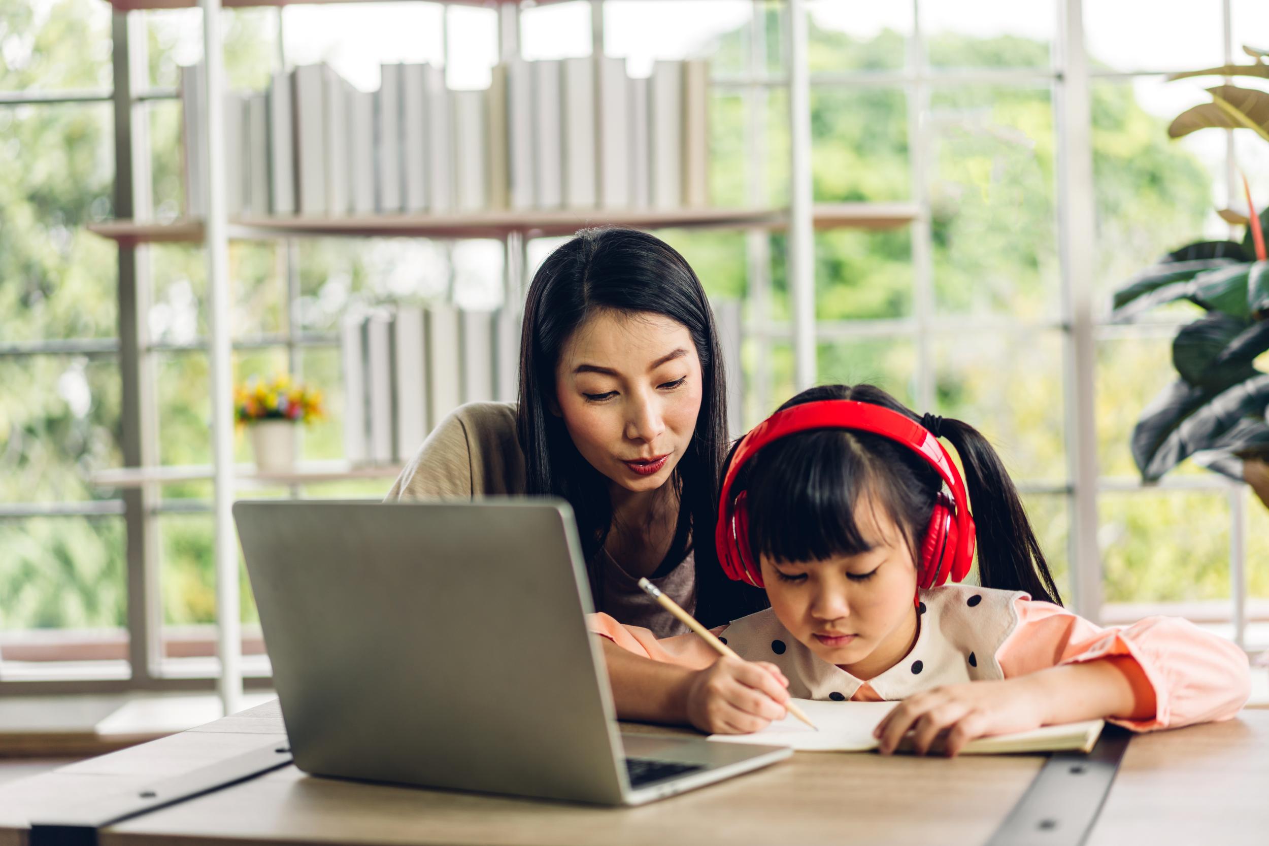 A woman and a girl wearing headphones work together at a table, with a laptop open and greenery