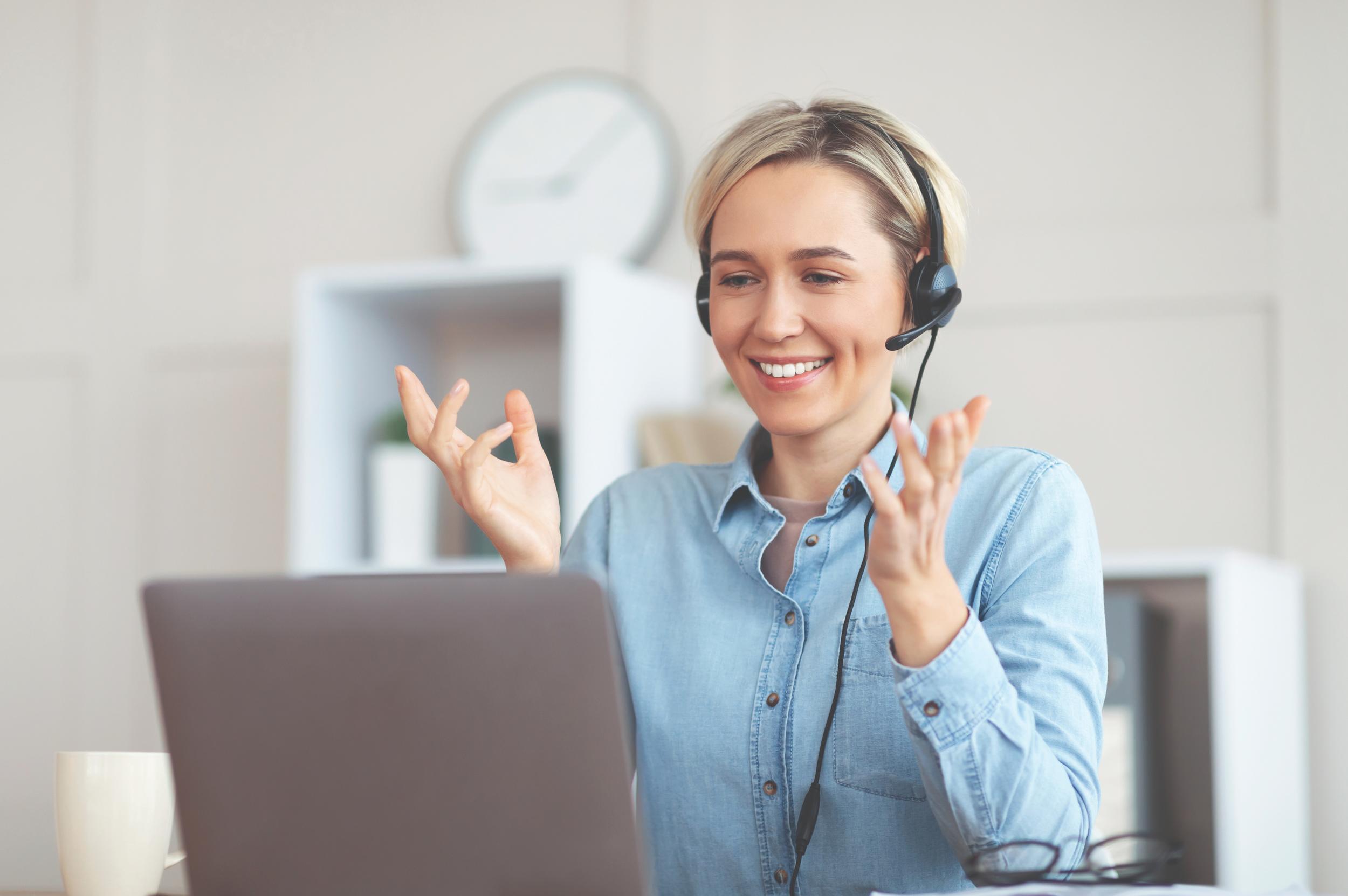 A woman wearing a headset smiles while engaging in a video call on her laptop, gesturing with her