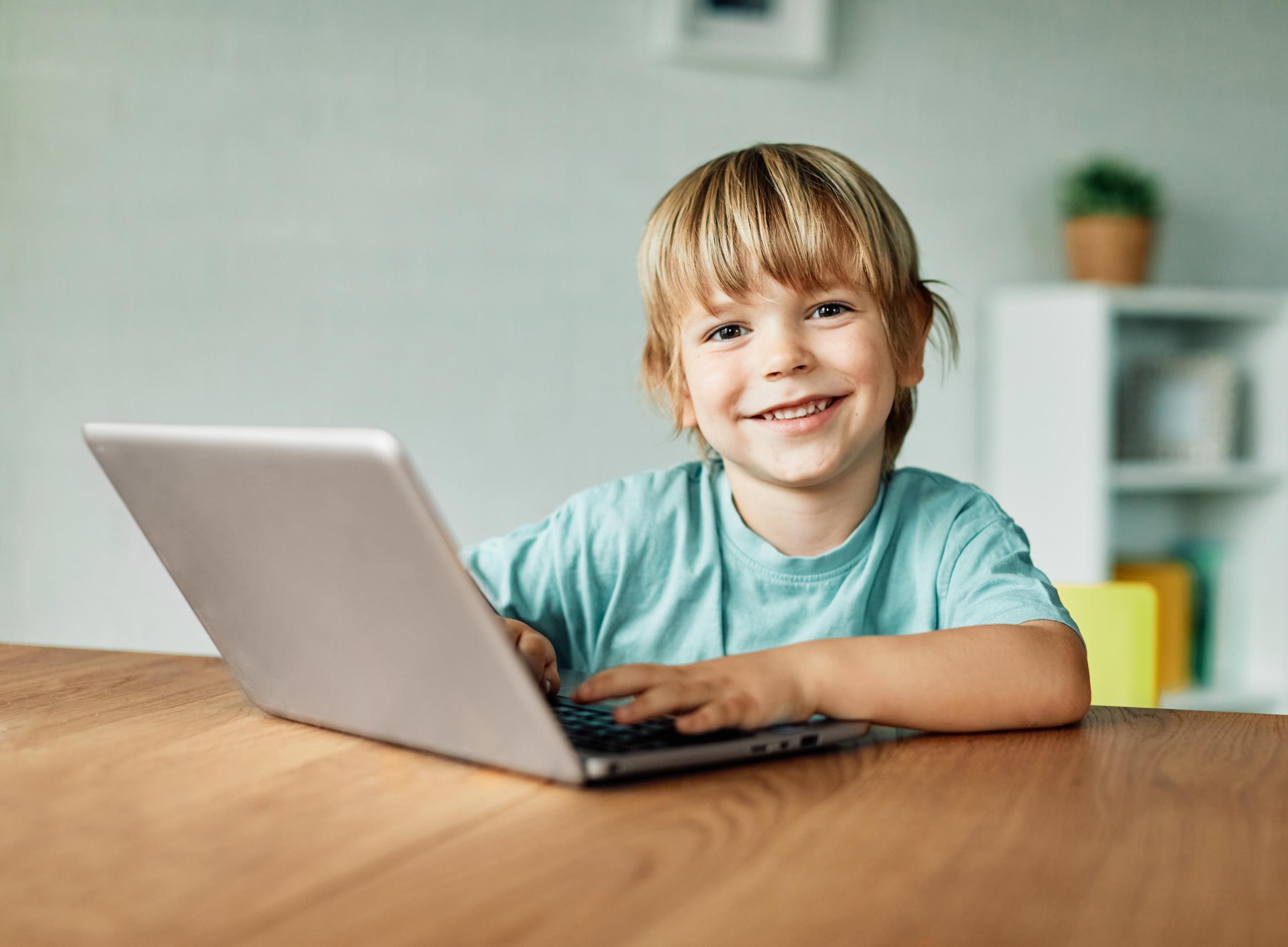 Young child with blonde hair smiles while sitting at a table, using a laptop. A bright, inviting room