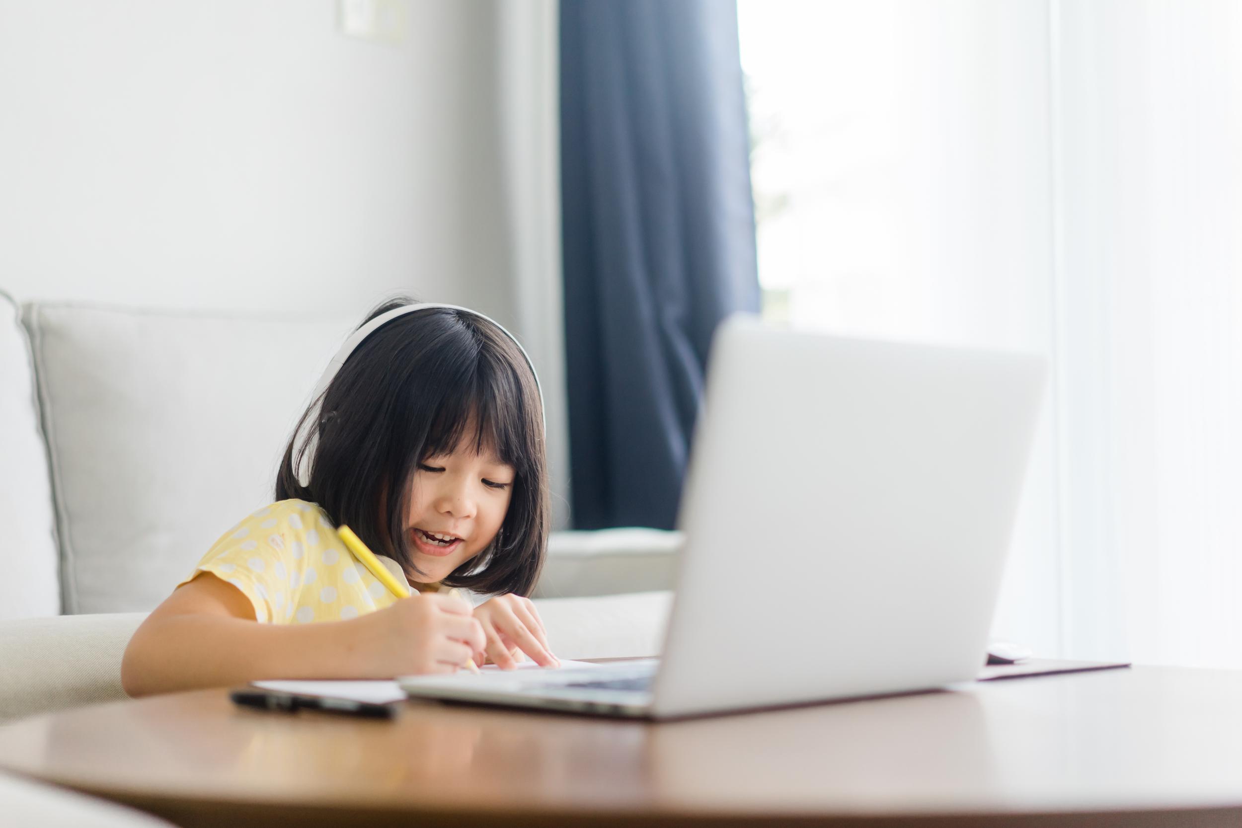 A young boy with headphones smiles while writing in a notebook next to a laptop on a table.