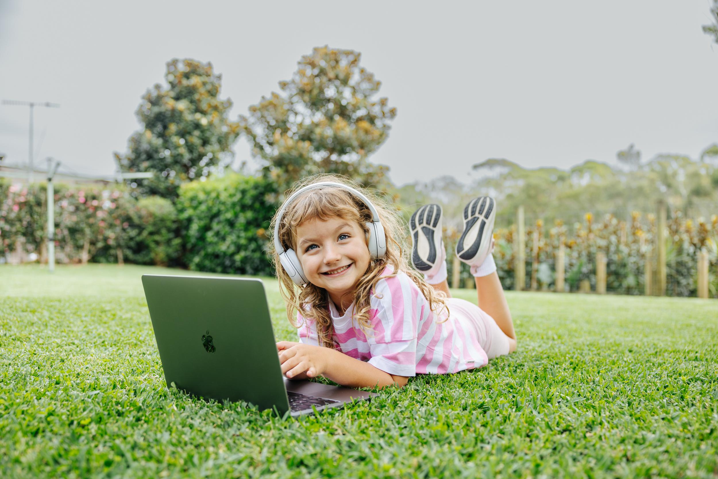A young girl lies on her stomach on grass, wearing headphones and using a laptop, smiling