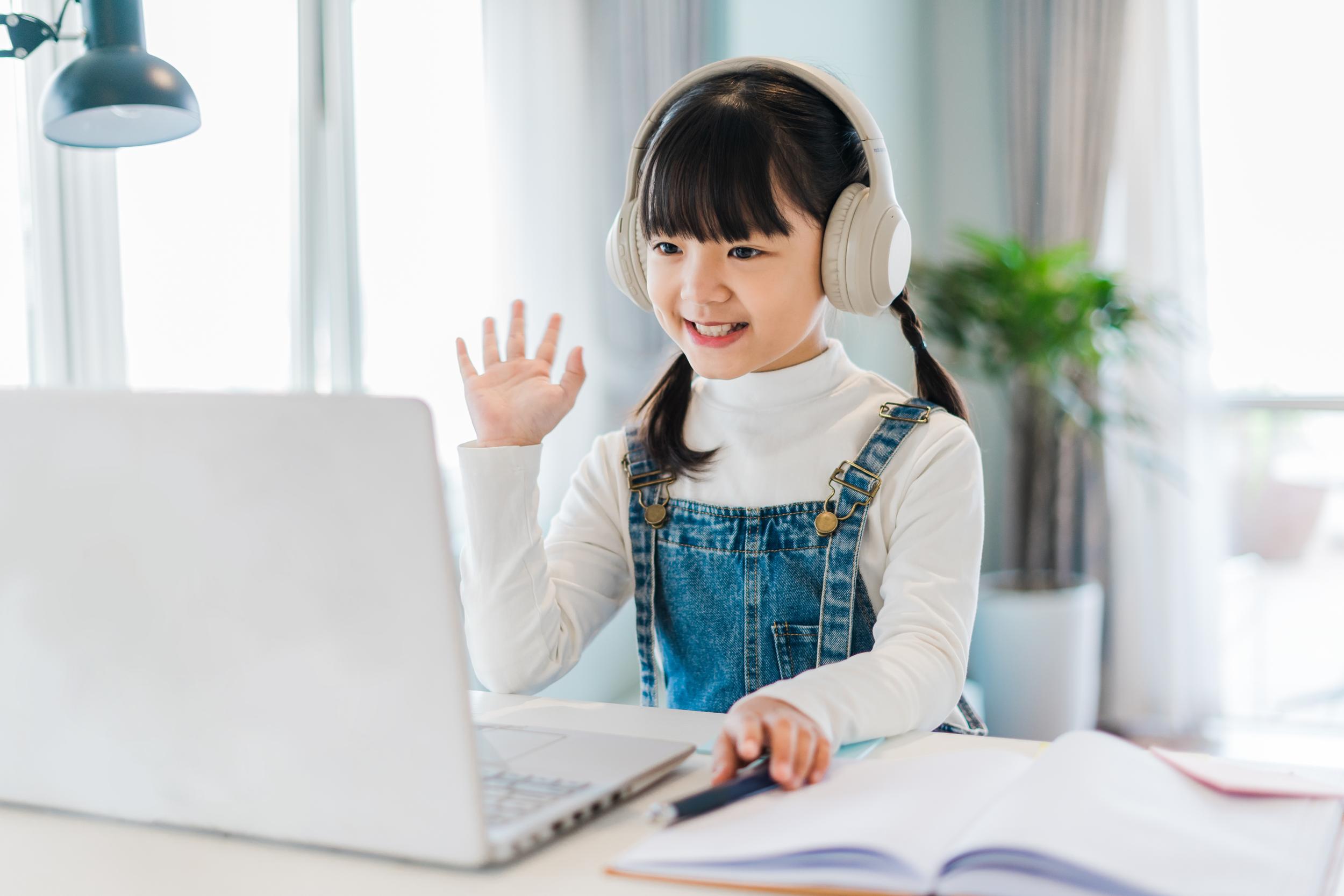 A young girl wearing headphones smiles and waves while sitting at a desk with a laptop, engaged