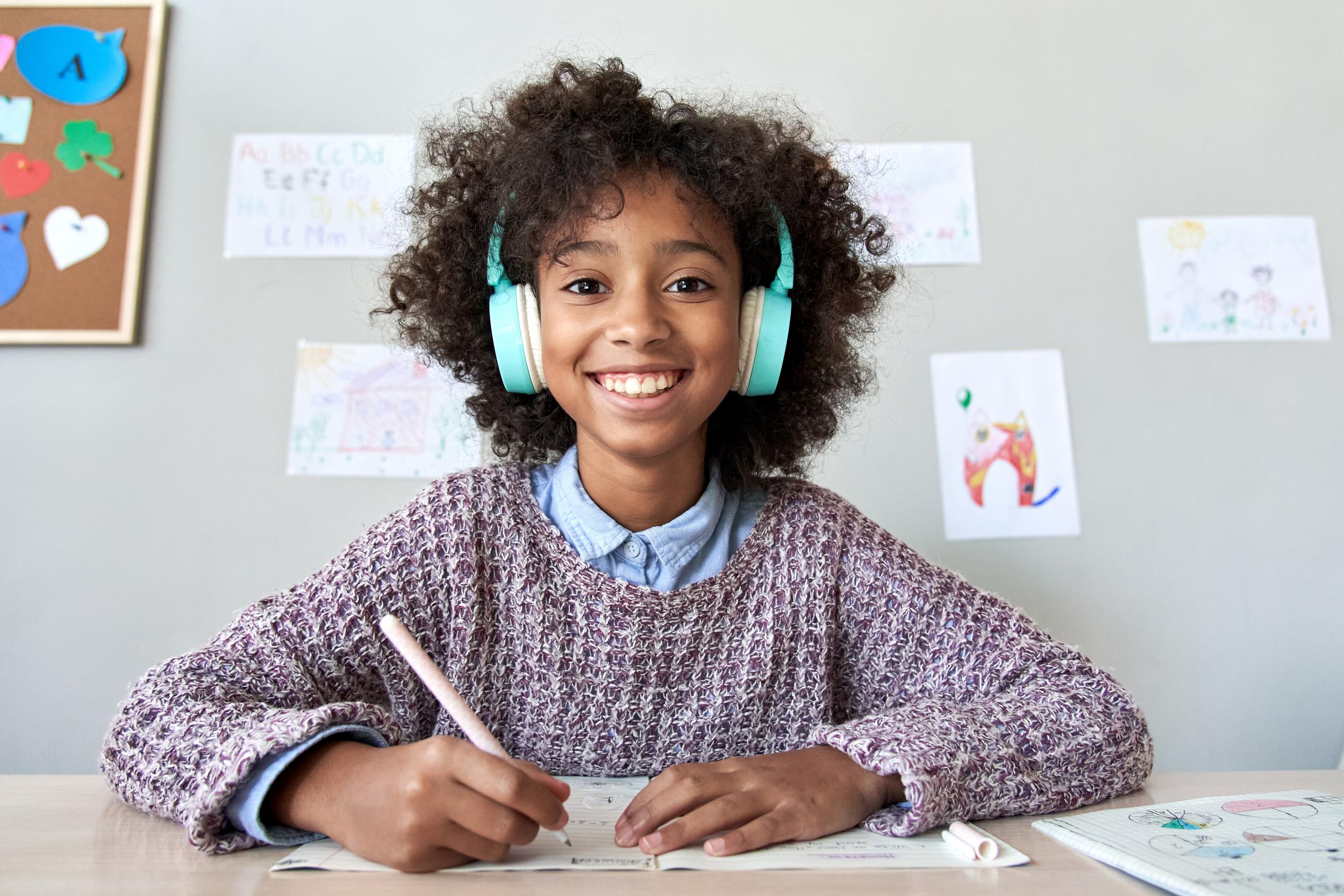 A young boy wearing headphones sits at a table, focused on a laptop, with a notebook and pen