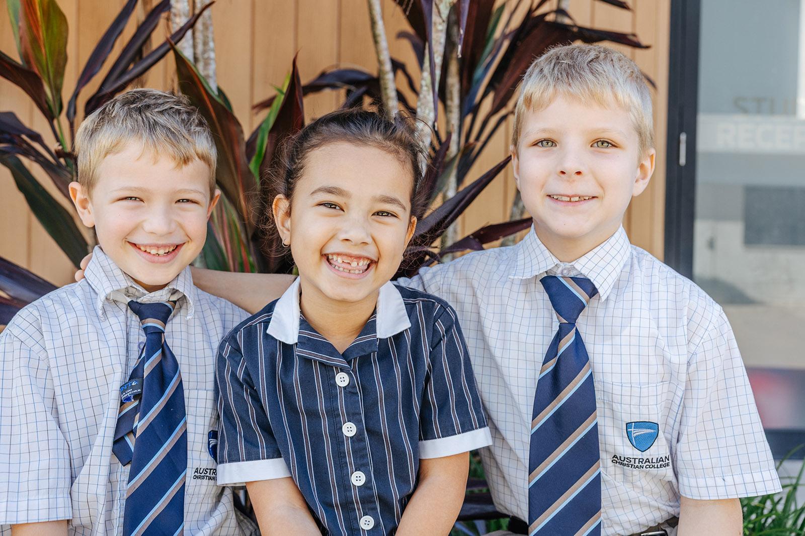 Three primary students smiling together outside the ACC Moreton school building