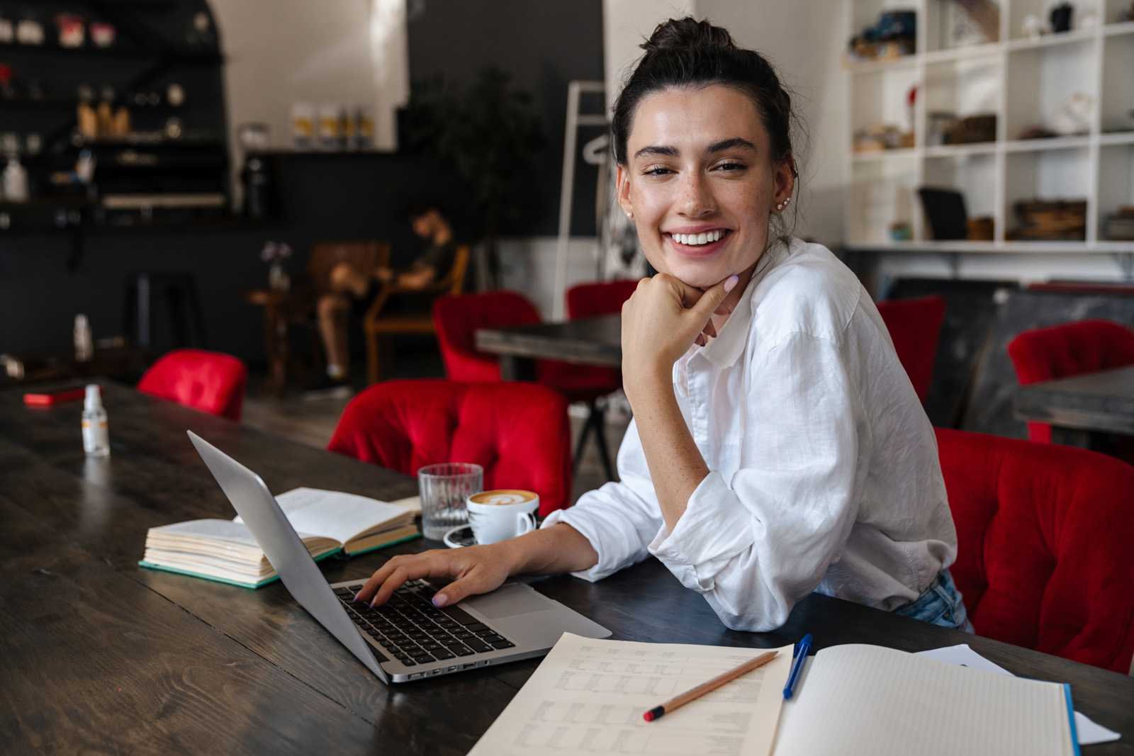 smiling older teenage girl sitting at table with a laptop, open books, and a cup of coffee