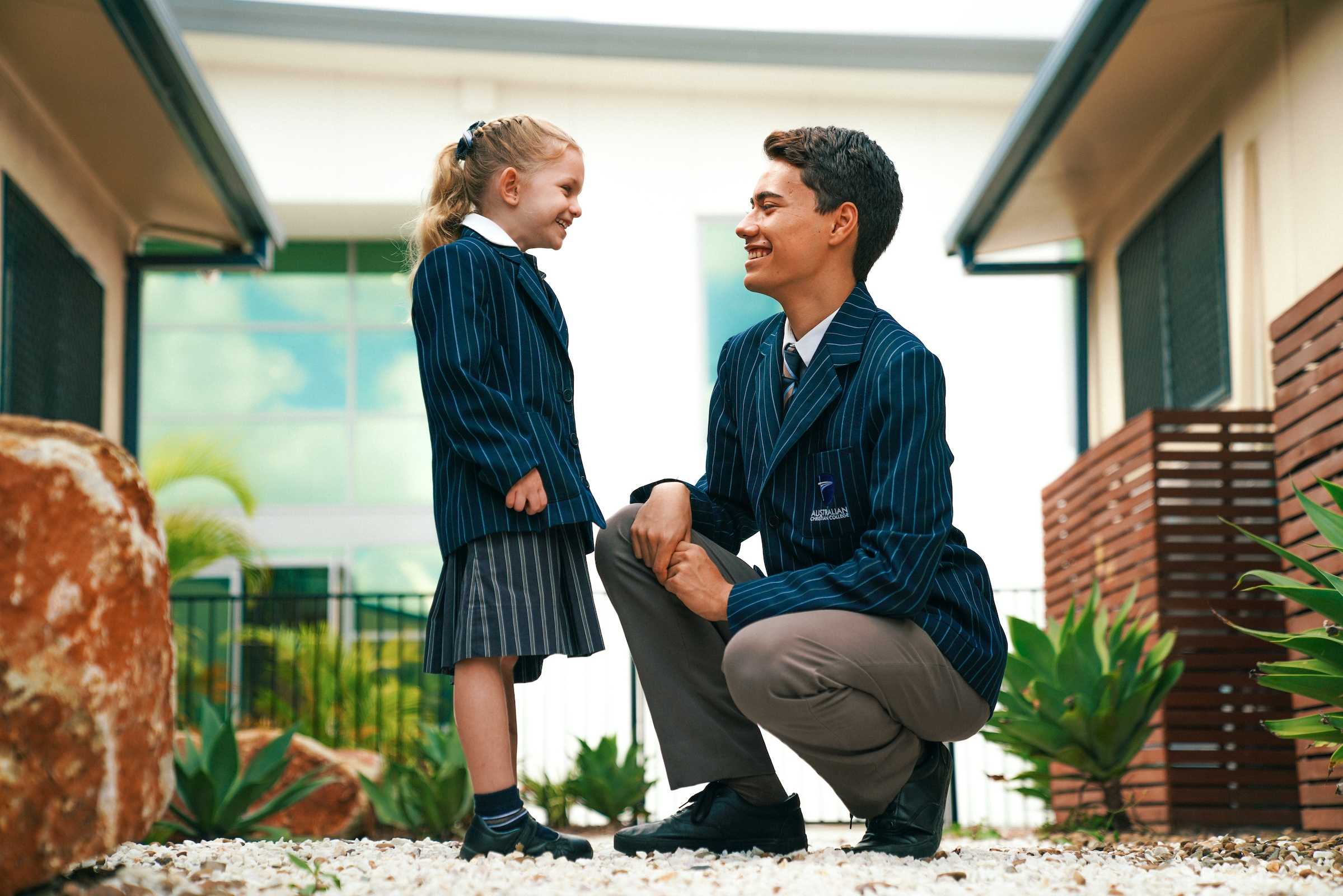 male high school student crouching next to young female primary school student, both smiling at each other