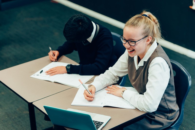 two students sitting at a desk writing in ther notebooks while laughing