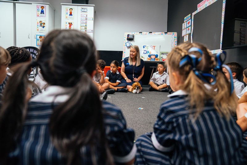 class of primary school students sitting in circle with teacher on floor of classroom