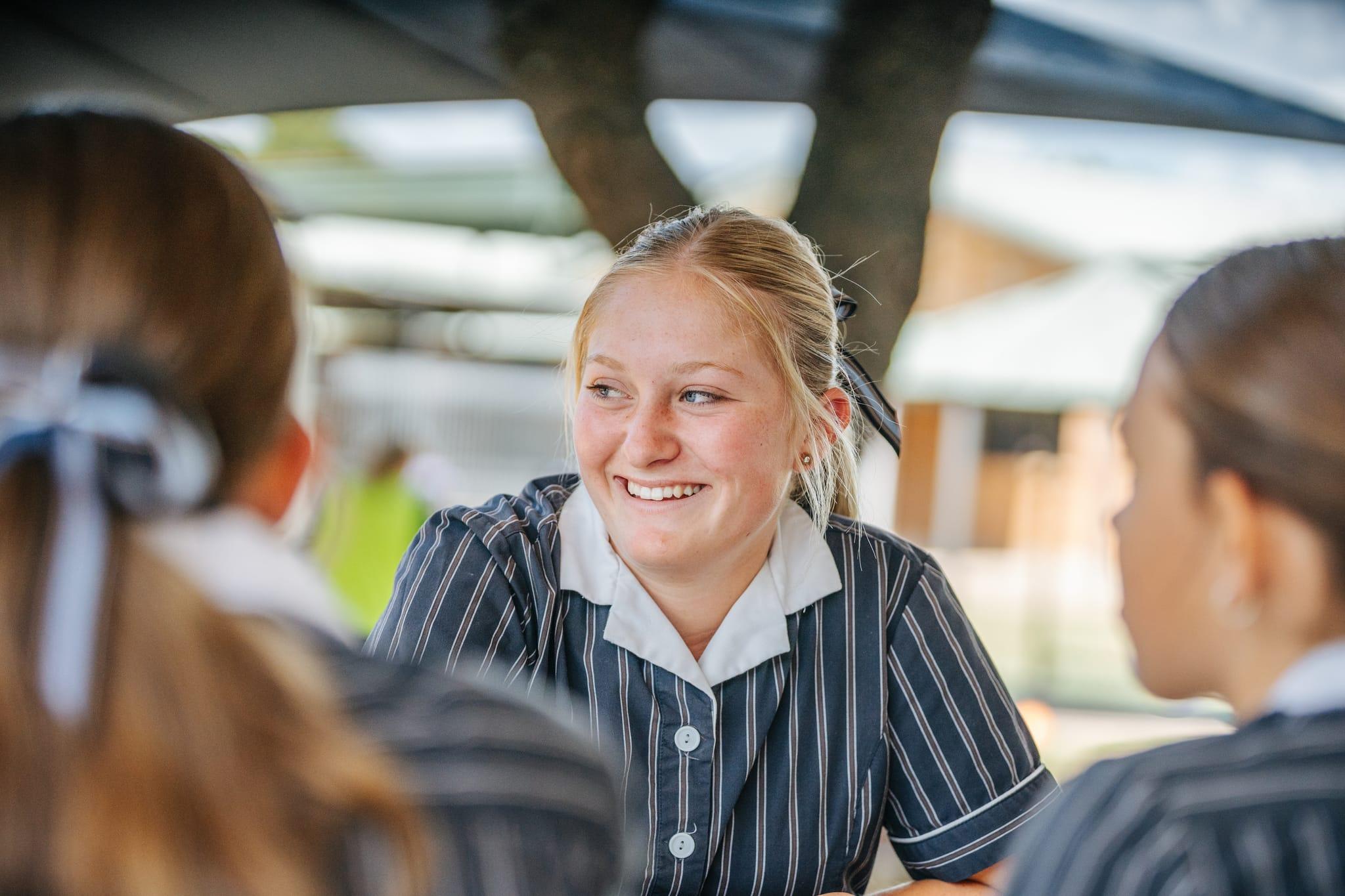 Smiling senior school girl in striped uniform chatting with friends outdoors.