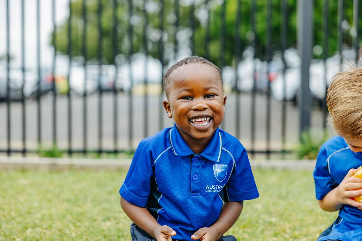A student at ACC Singleton smiles while sitting on the grass, with a fence and cars visible