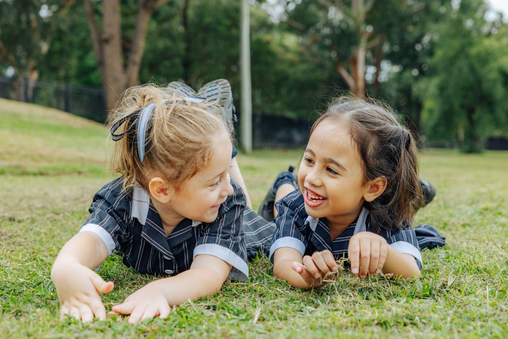 Two junior school girls smiling and talking on the grass at ACC Singleton, capturing genuine connection and student belonging