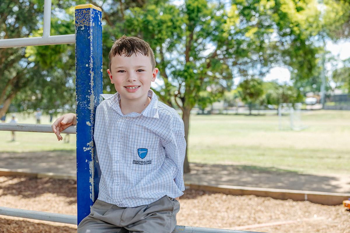 A student in a school uniform sits on playground equipment at ACC Singleton, smiling in a sunny outdoor