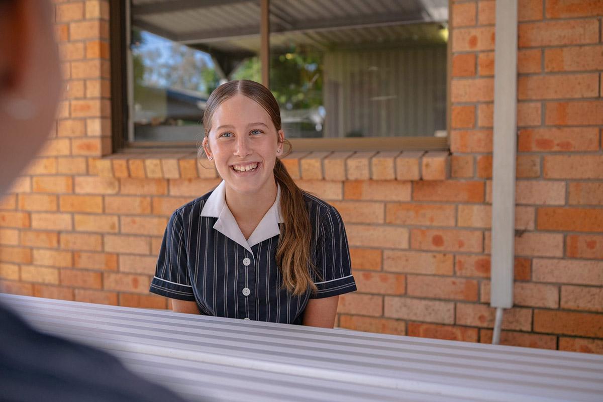 A student in a school uniform smiles while seated at a table outside ACC Singleton, with a brick