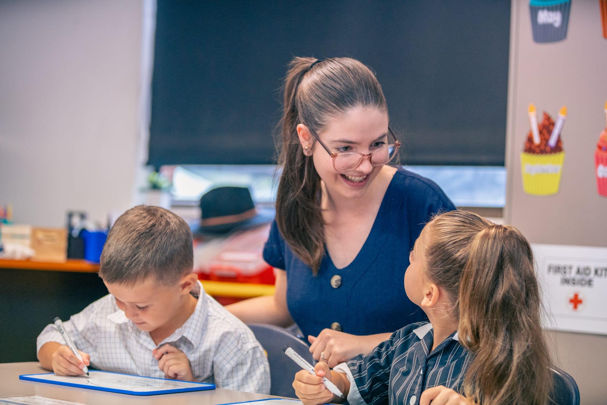 Friendly junior school teacher engaging with young students during a classroom learning activity at a Christian school.