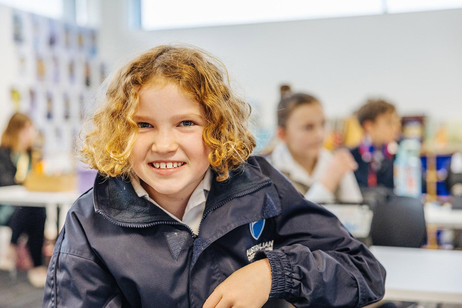 Smiling primary girl in ACC jacket inside an Albany classroom with classmates seated behind her