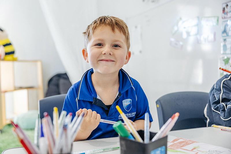 Smiling kindergarten boy at a classroom desk with coloured pencils refining fine motor skills through drawing