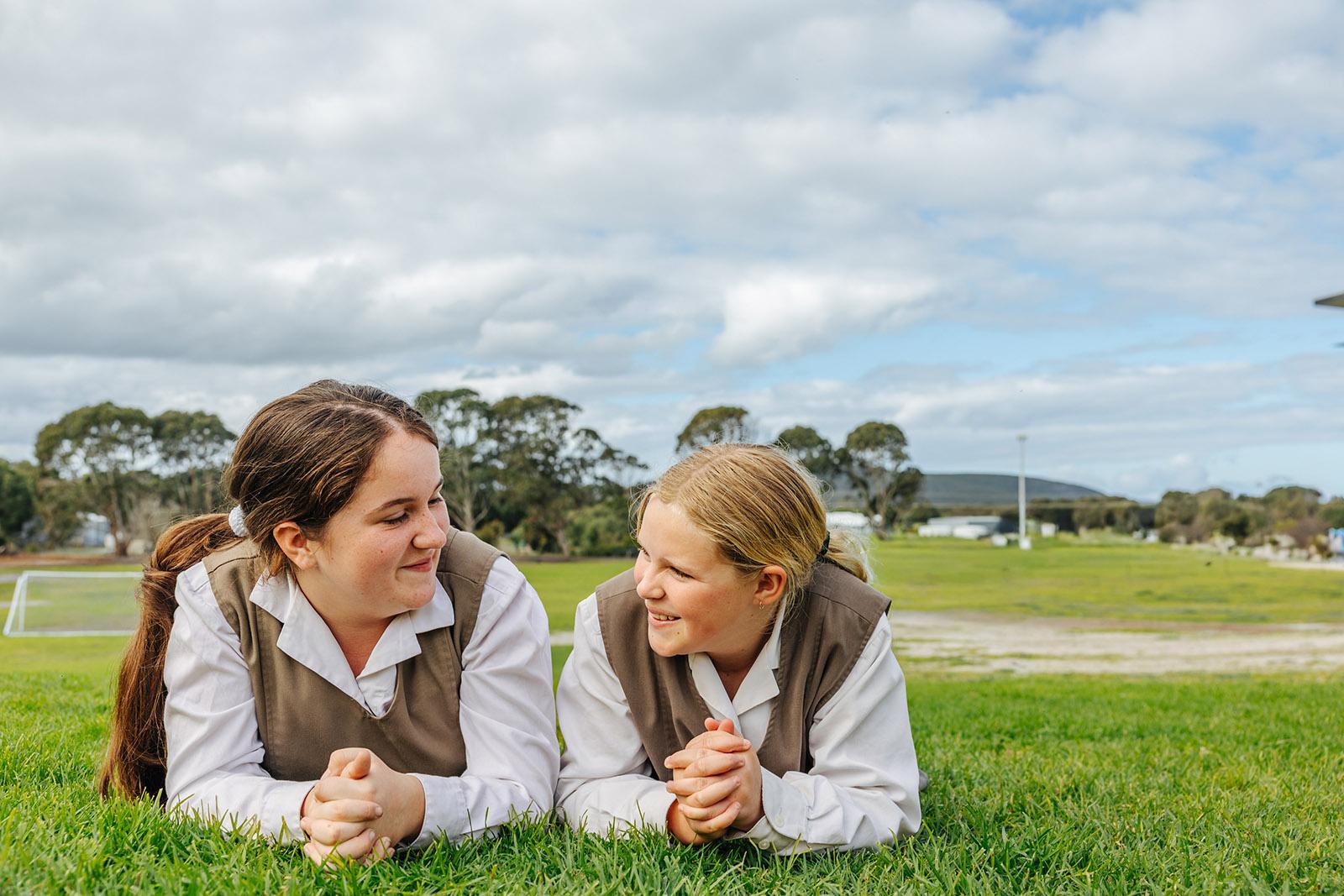 Two primary girls in ACC Southlands formal uniforms lying on the school grass laughing together
