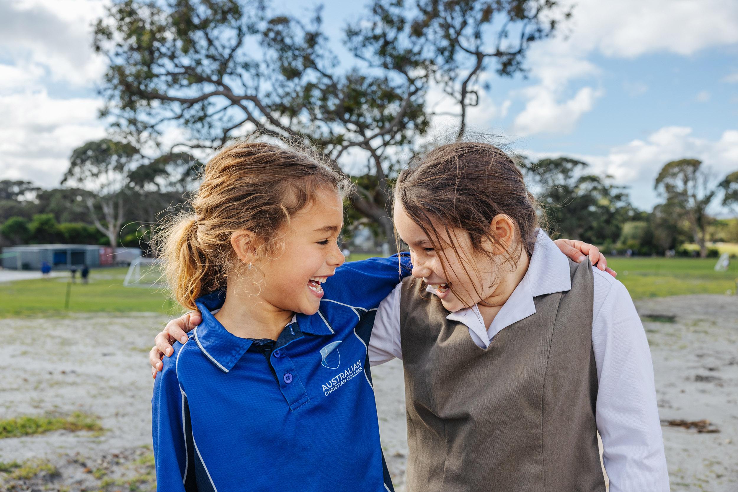 Two primary girls in ACC Southlands uniforms laughing together outdoors on school grounds