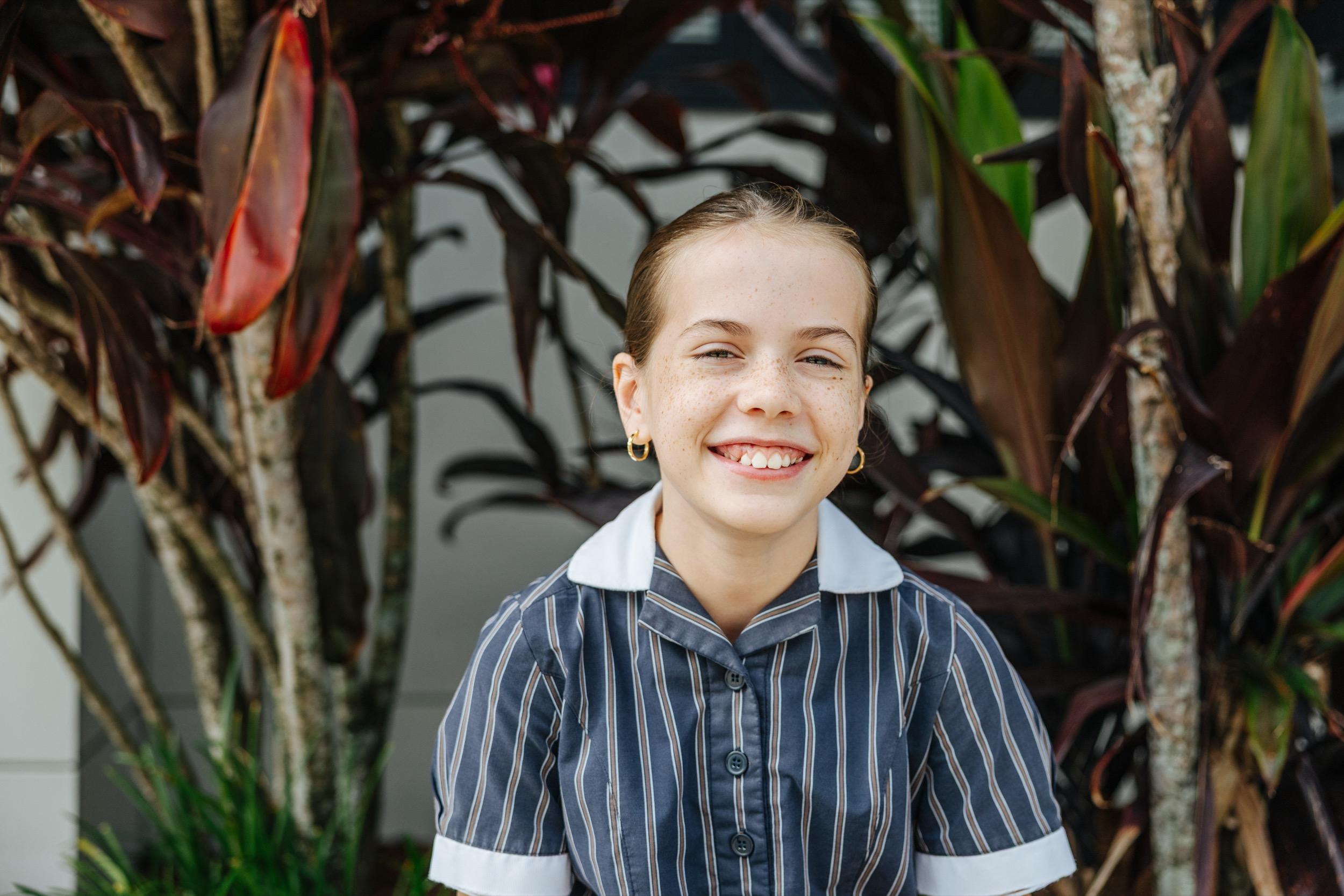A student in school uniform smiles while sitting in front of vibrant plants, a friendly expression.