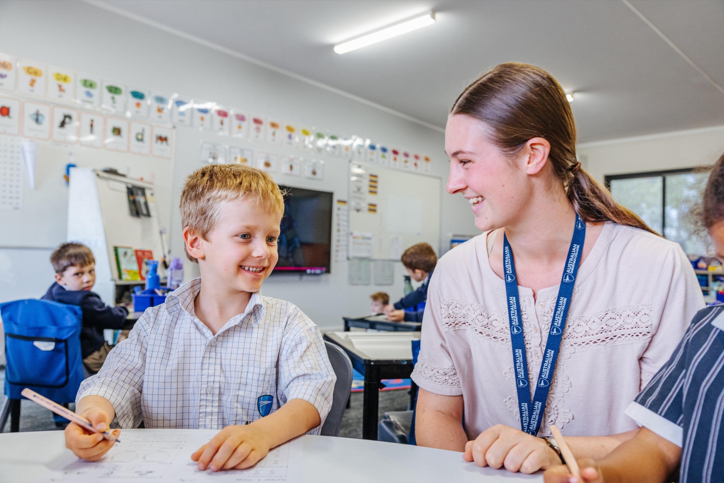 A teacher and a young student smile at each other while working on a worksheet at a classroom