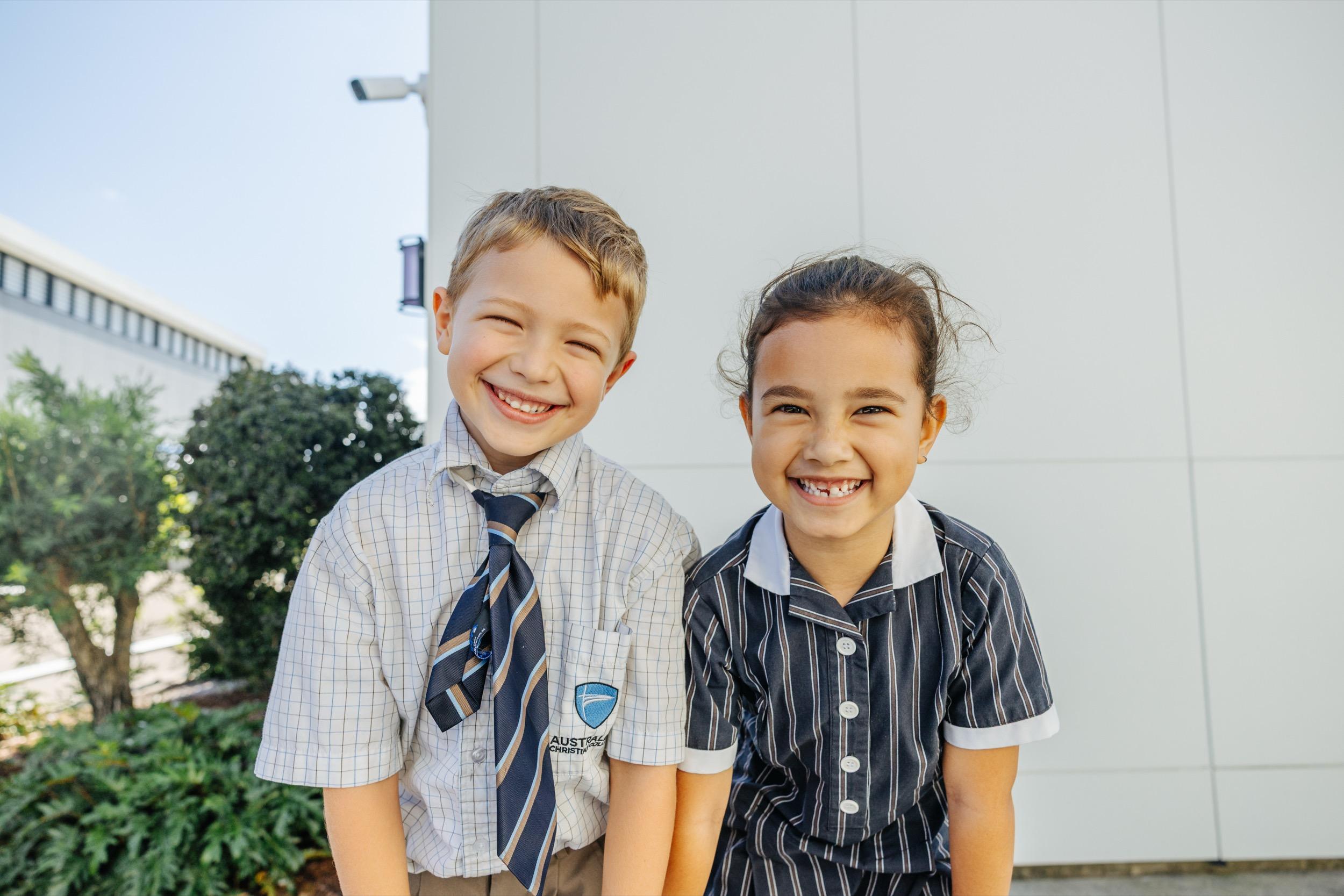 Two smiling students in school uniforms stand together outside ACC Swan Hill, with greenery and a building