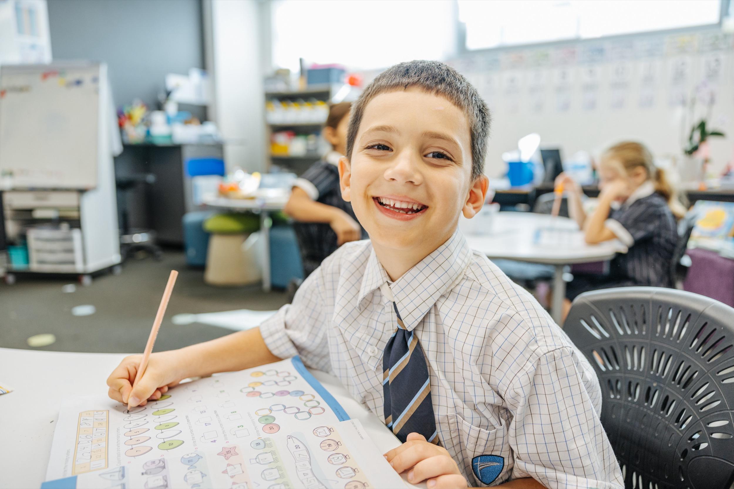 A smiling student in a school uniform sits at a desk, working on a colorful worksheet