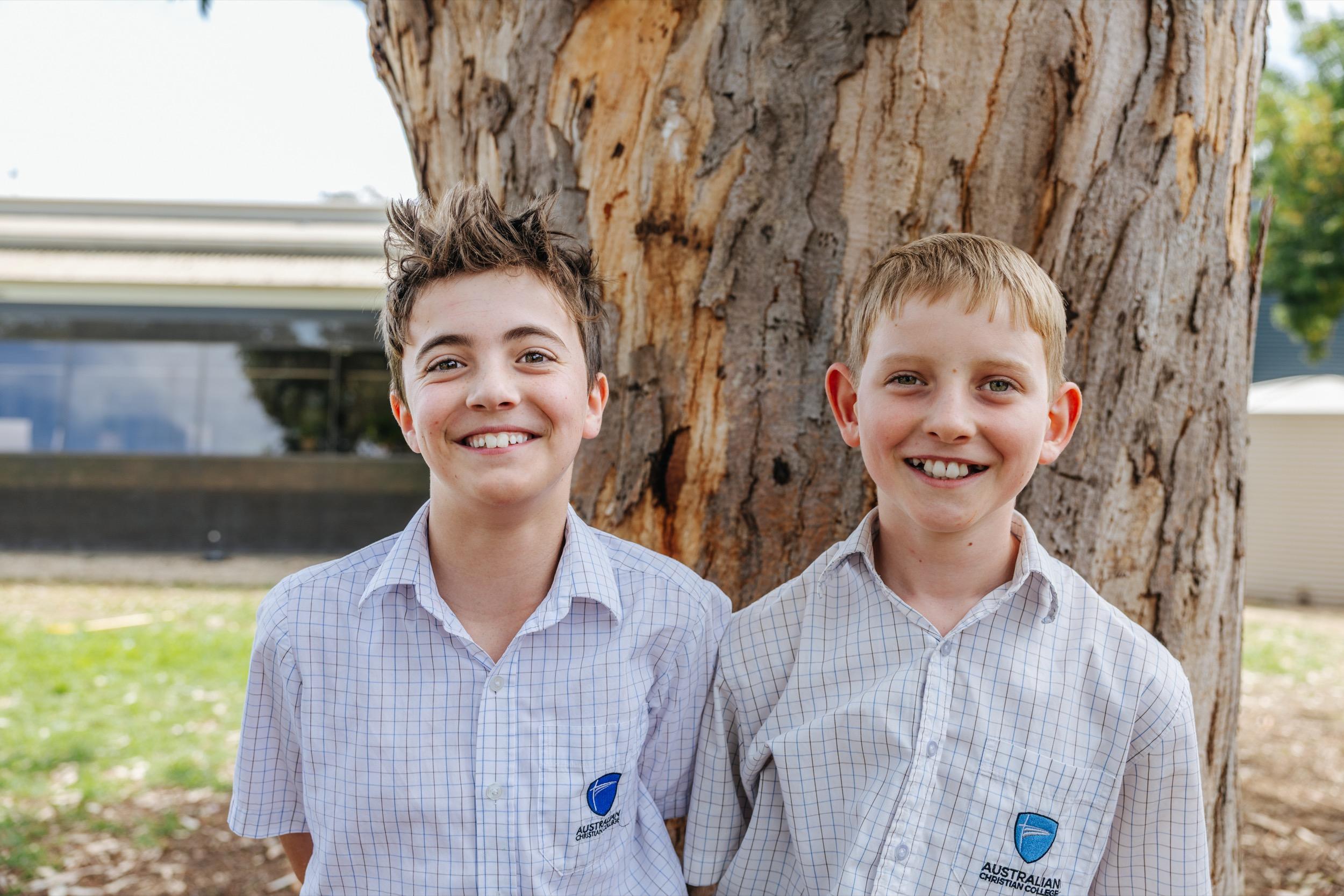 Two students in school uniforms smiling together, standing beside a large tree at ACC Swan Hill.