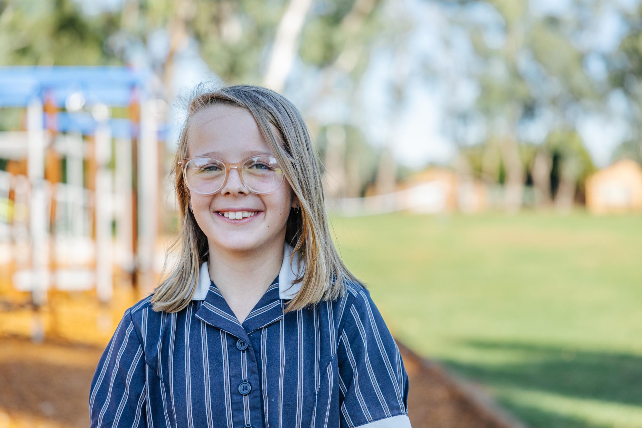 Smiling student in school uniform stands in playground at ACC Swan Hill, with play equipment and green grass