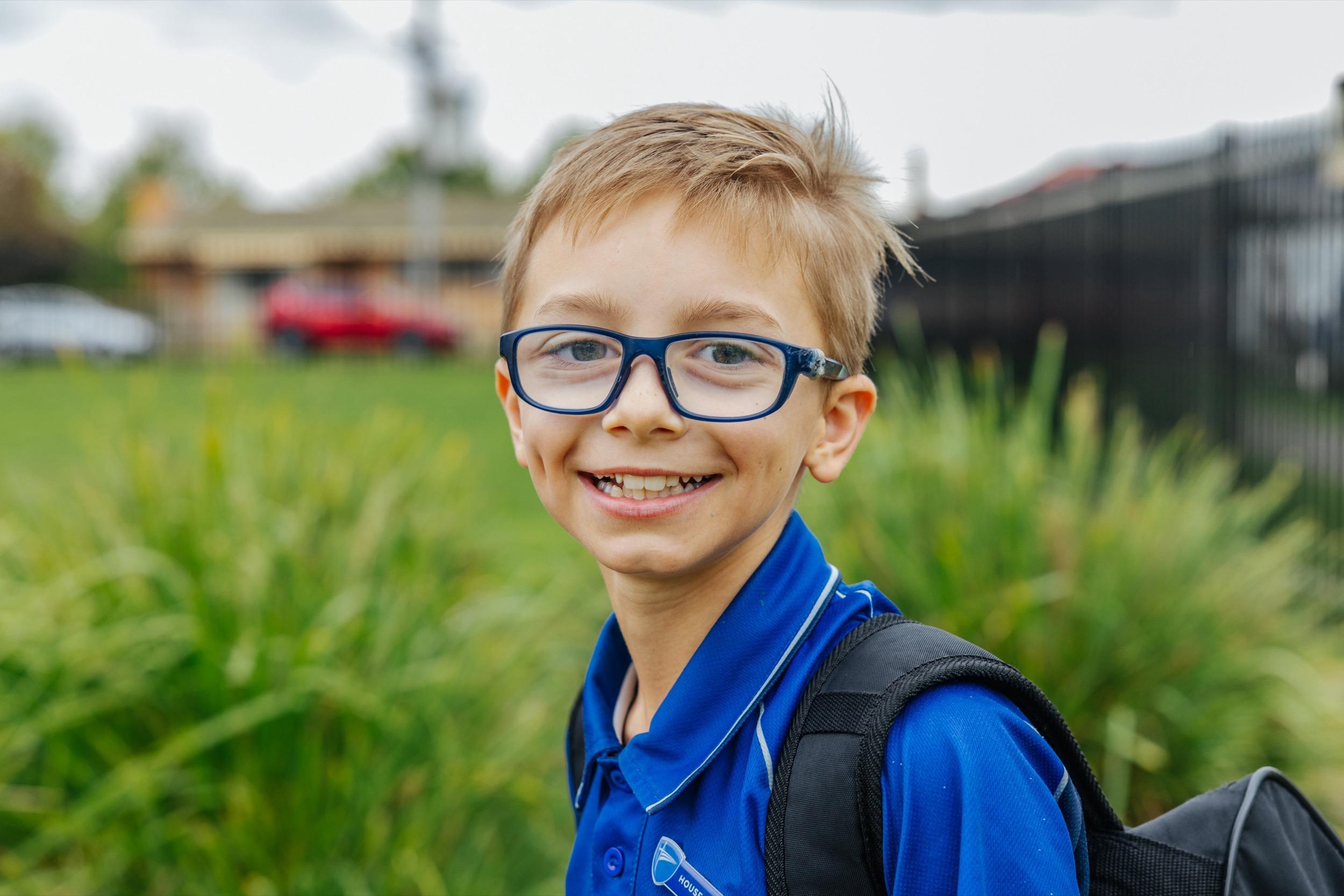 Smiling student wearing a school uniform and glasses stands outdoors, with greenery and buildings in the background.