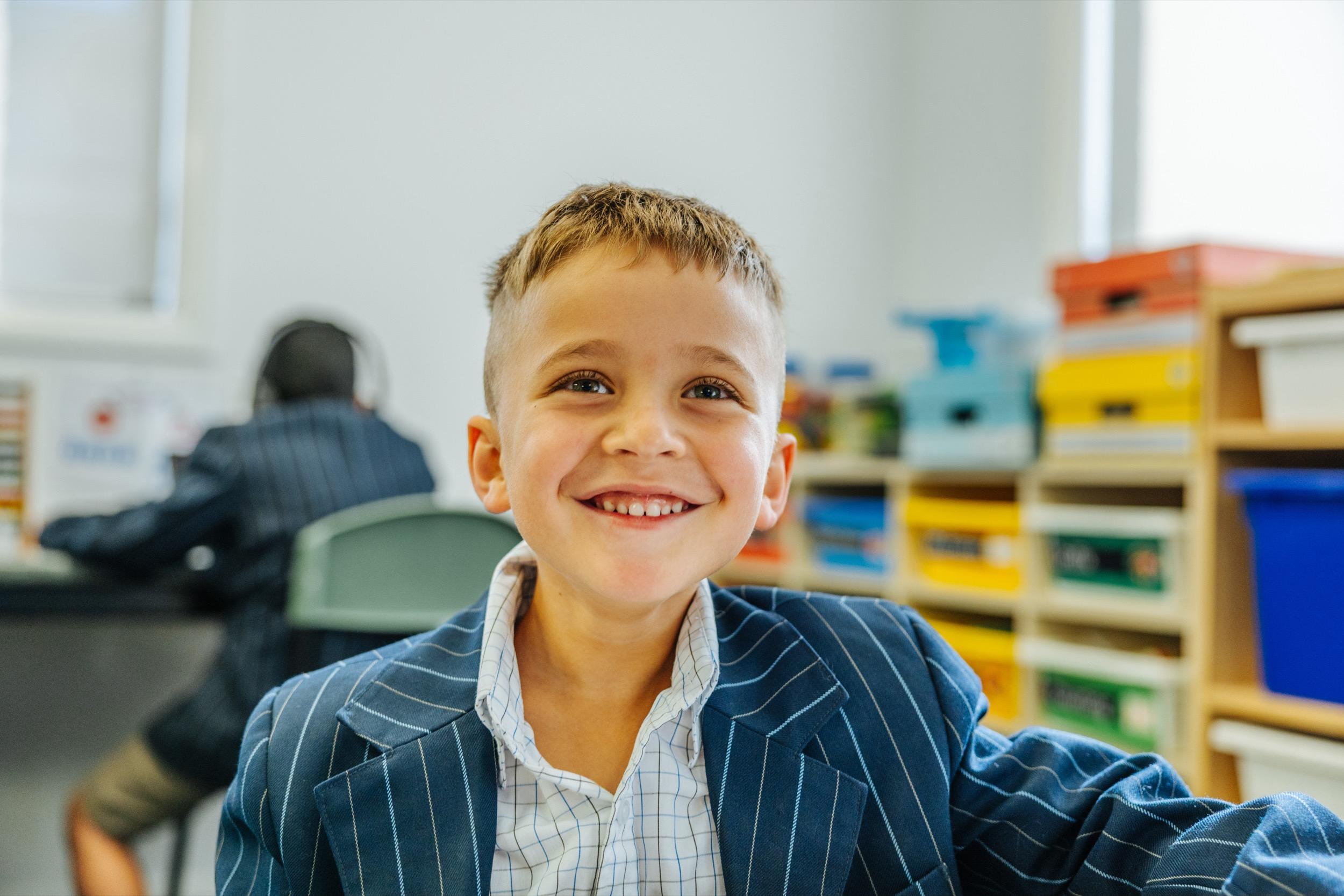A smiling boy in a school uniform sits at a desk, with colorful storage boxes and another student
