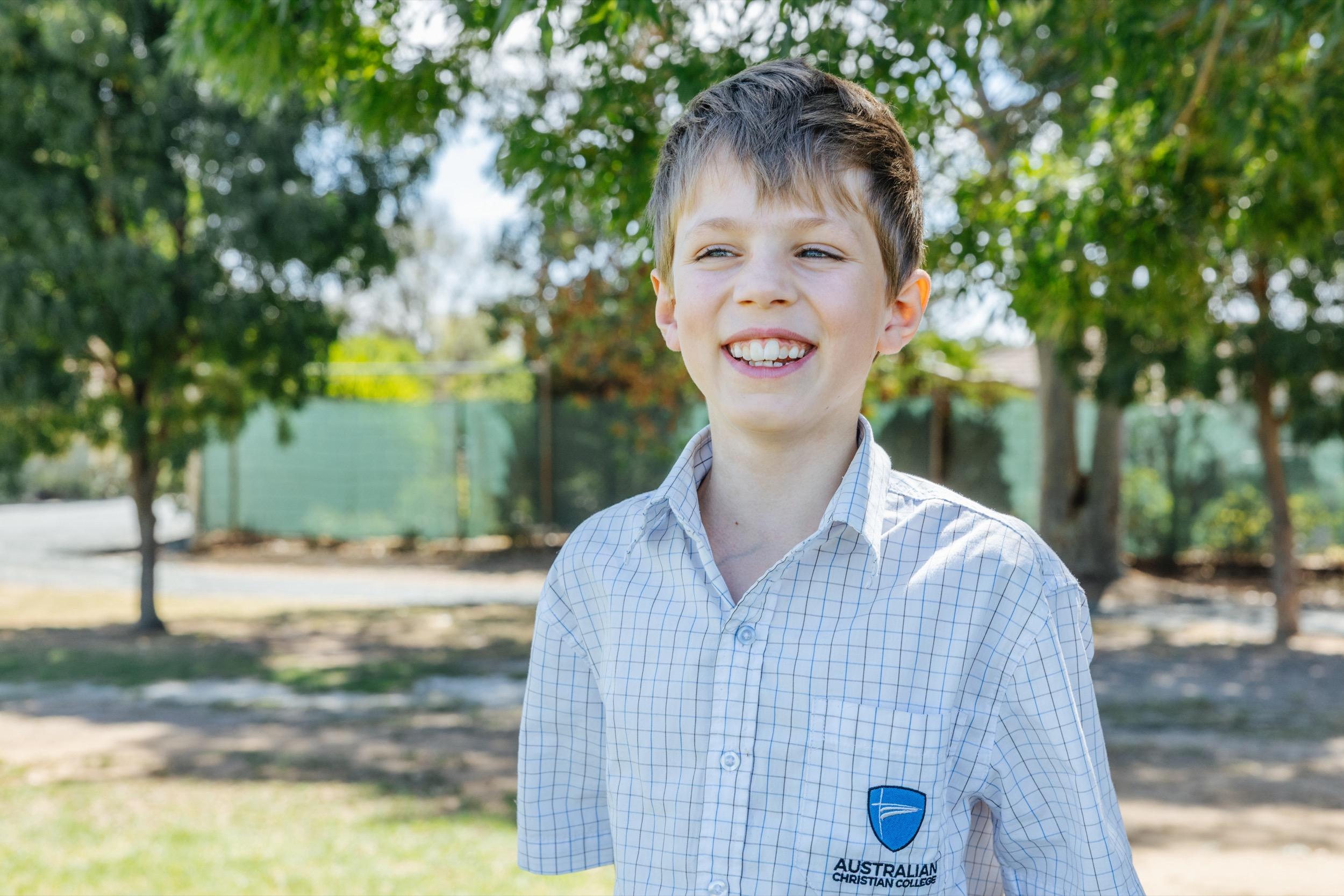 A student in a school uniform smiles while standing outdoors among trees, enjoying a sunny day.