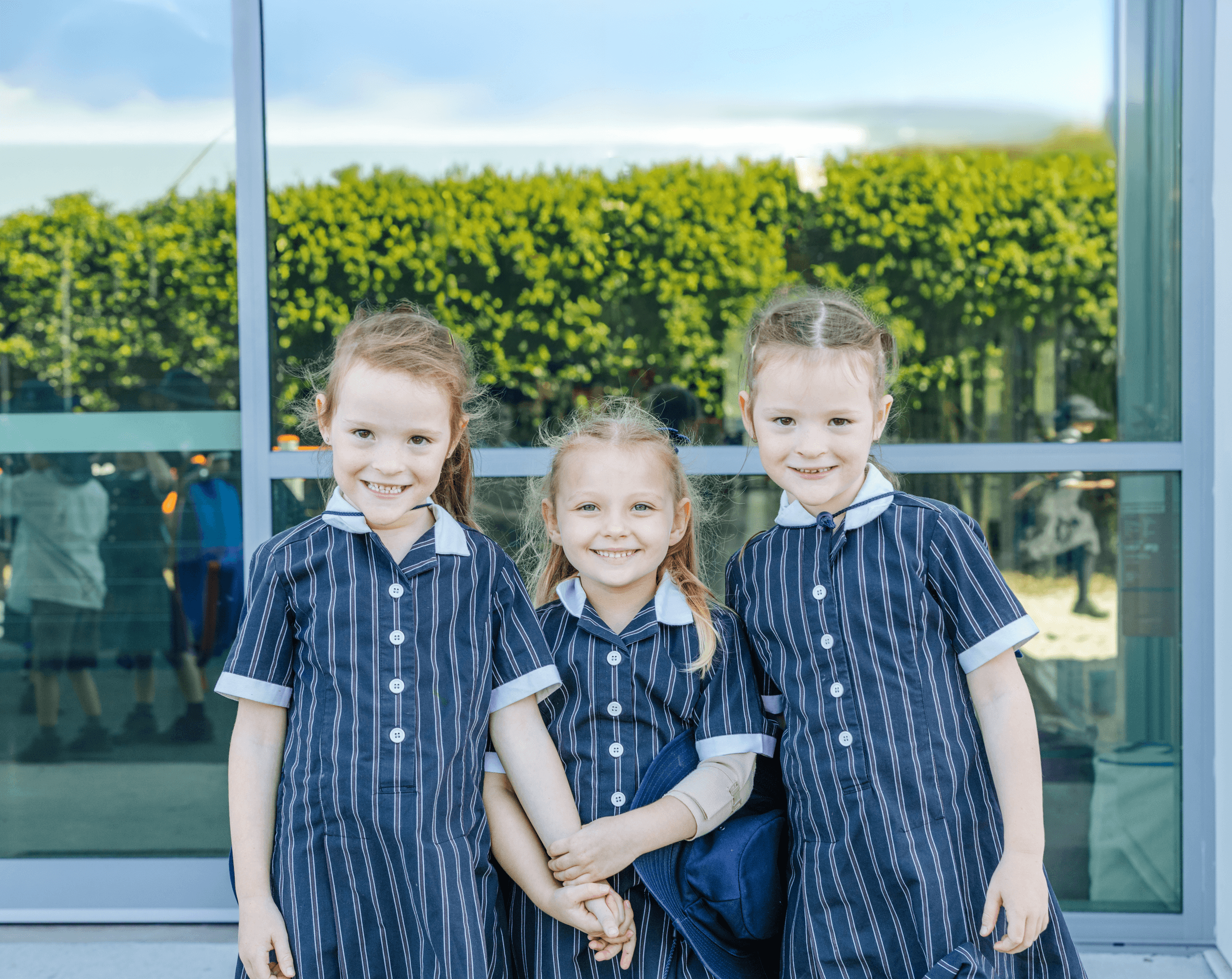 Three students in school uniforms stand together, smiling in front of a glass wall with greenery reflected.